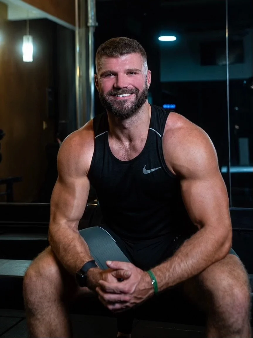 A muscular man with short hair and a beard, smiling and sitting in a gym. He's wearing a black sleeveless athletic shirt, black shorts, and a watch, with a fitness tracker on his ankle.