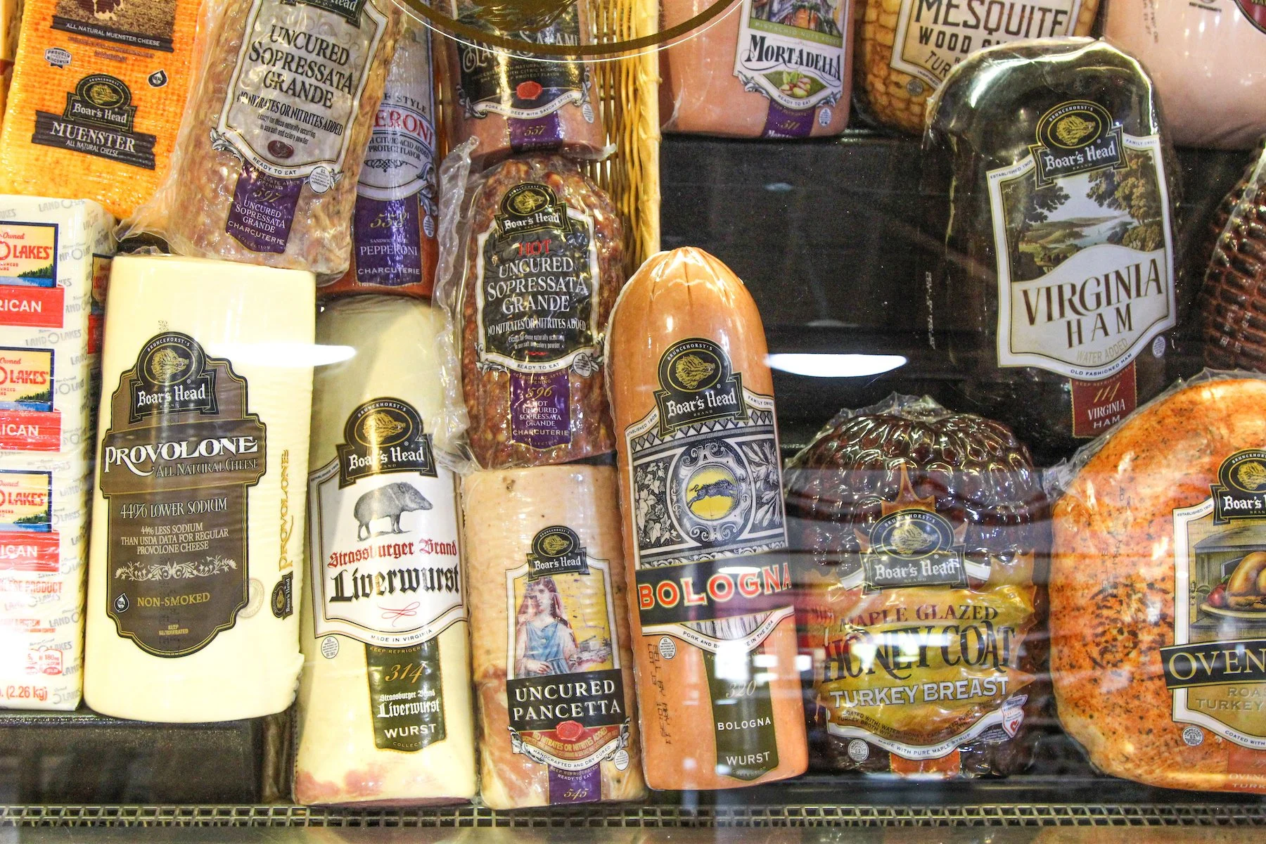 A display case filled with various types of meats and cheeses. Visible are packages of Bologna, Virginia Ham, Liverwurst, uncured Sopressata, pancetta, honey coat turkey breast, and slices of provolone cheese. Some packages have labels with brand nam
