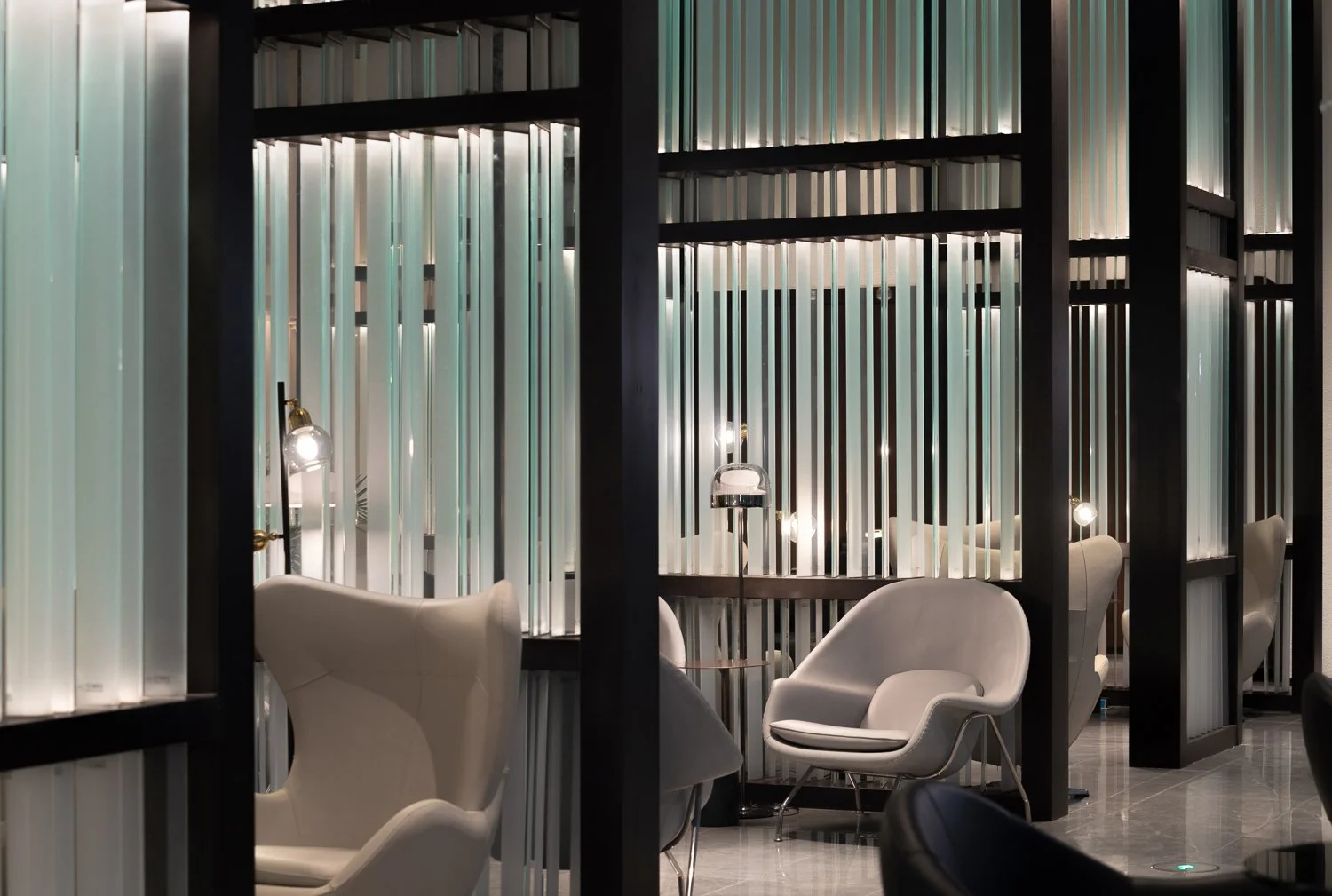 Modern lounge area with white armchairs, black partition walls with vertical striped panels, small lamps, and marble flooring.