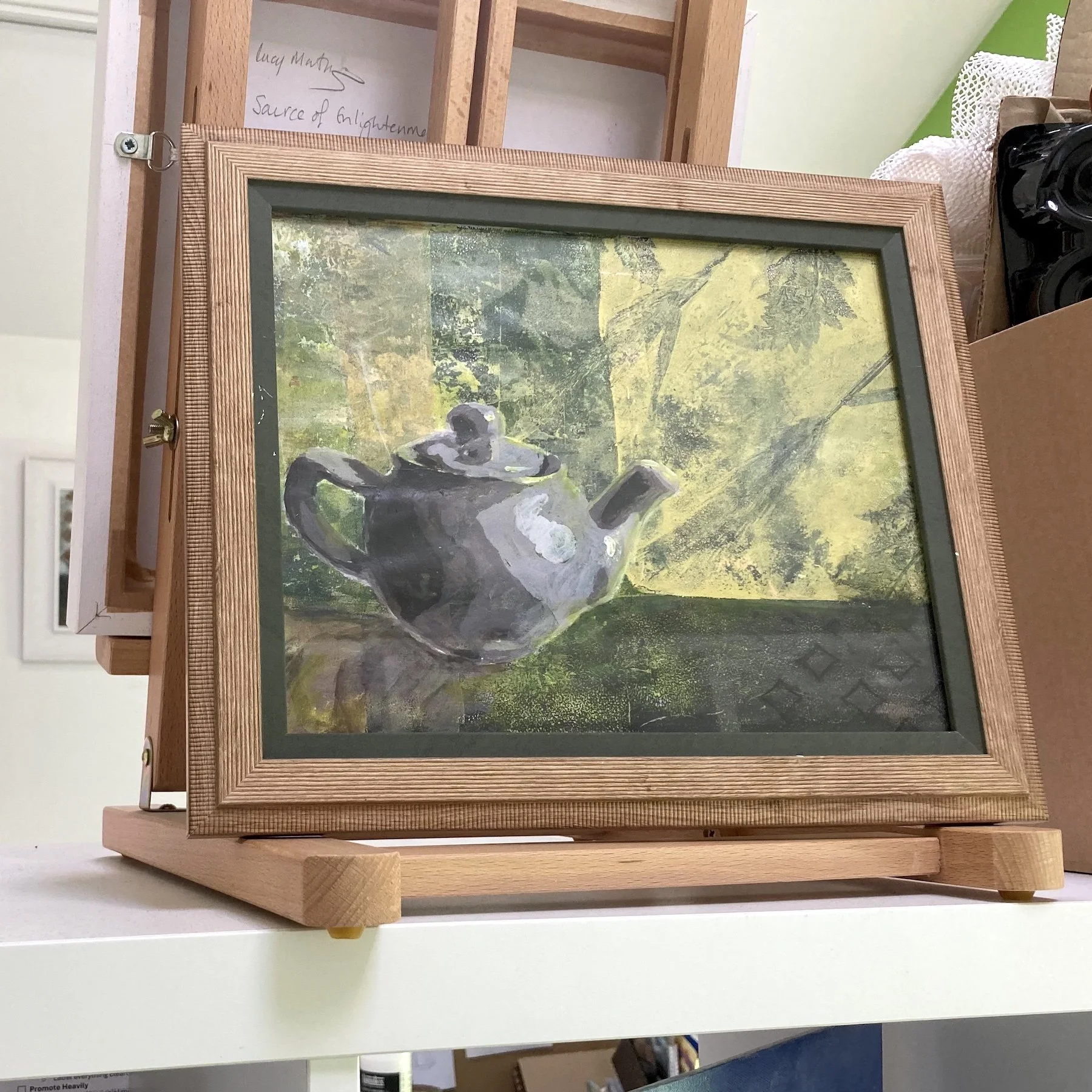 Lucy Mathers' painting called Green Tea shown in the studio, framed in a wood-effect frame with dark green insert.