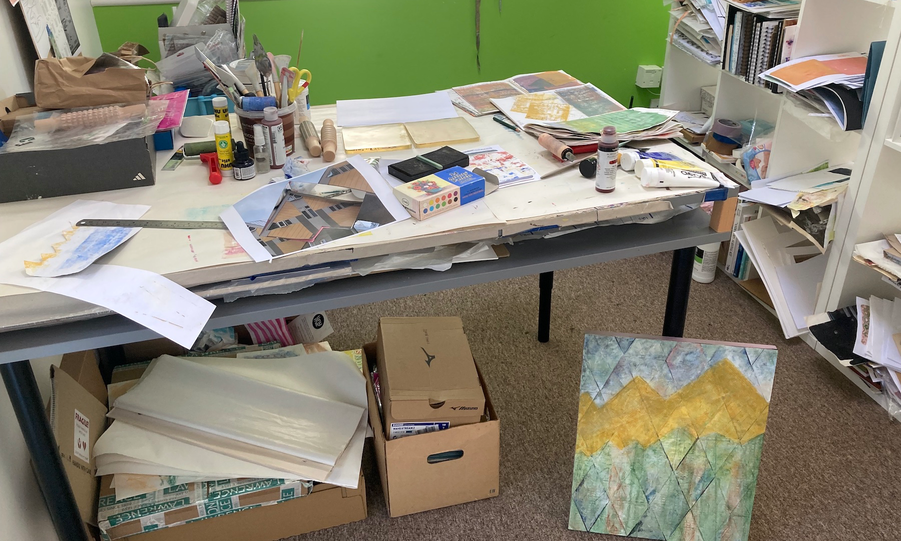 A view of Lucy Mathers Studio showing her art table with various art-making materials in tubs. There are pages of papers, soft pastels, paint, two gel plates, a printed photograph and a metal ruler on the desk and a painting leaning against it.