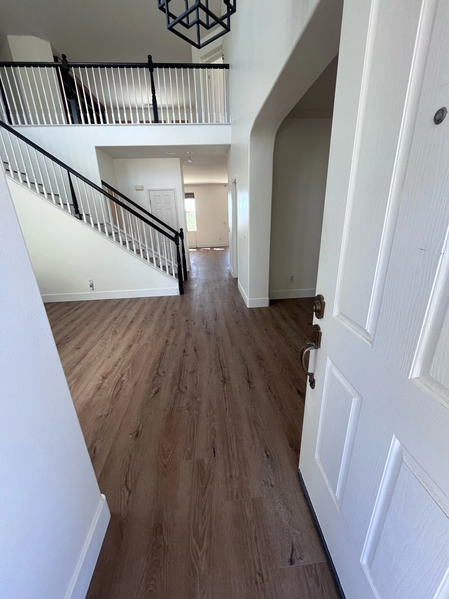 View from an open door into a modern multi-level house interior with a wooden floor, white walls, staircase with black railing, and upper balcony.