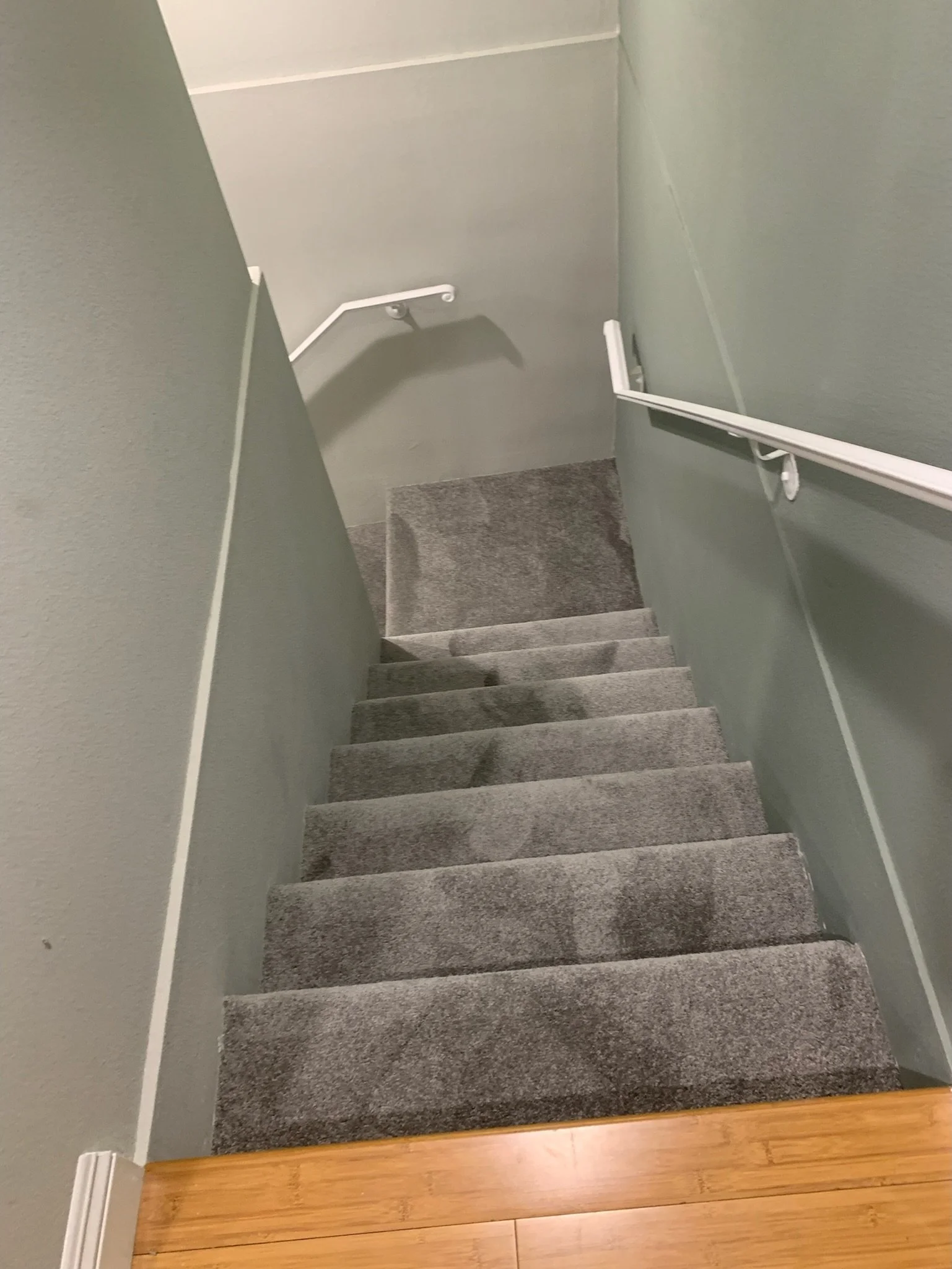 Top-down view of a staircase with gray carpeted steps, beige wooden flooring at the top, light green walls, and white handrails on both sides.