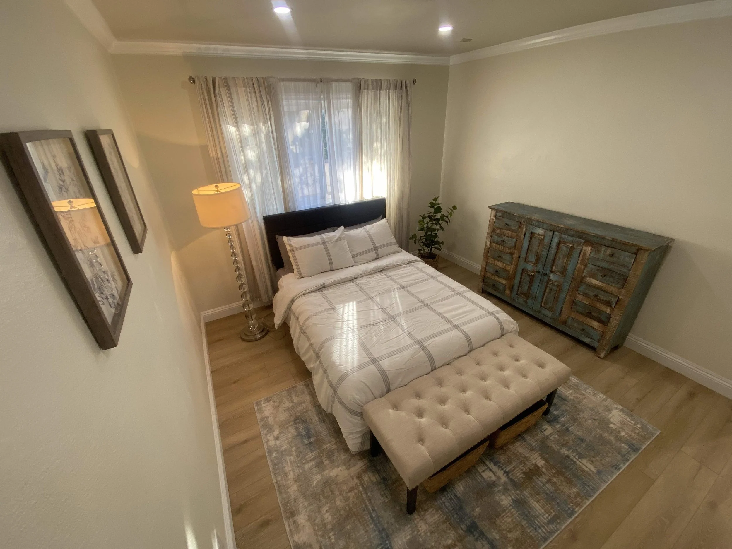 A cozy bedroom with a bed covered in white and gray plaid bedding, a beige tufted bench at the foot, a rustic blue dresser on the right, a tall floor lamp on the left, a window with sheer curtains behind the bed, a potted plant in the corner, and fra