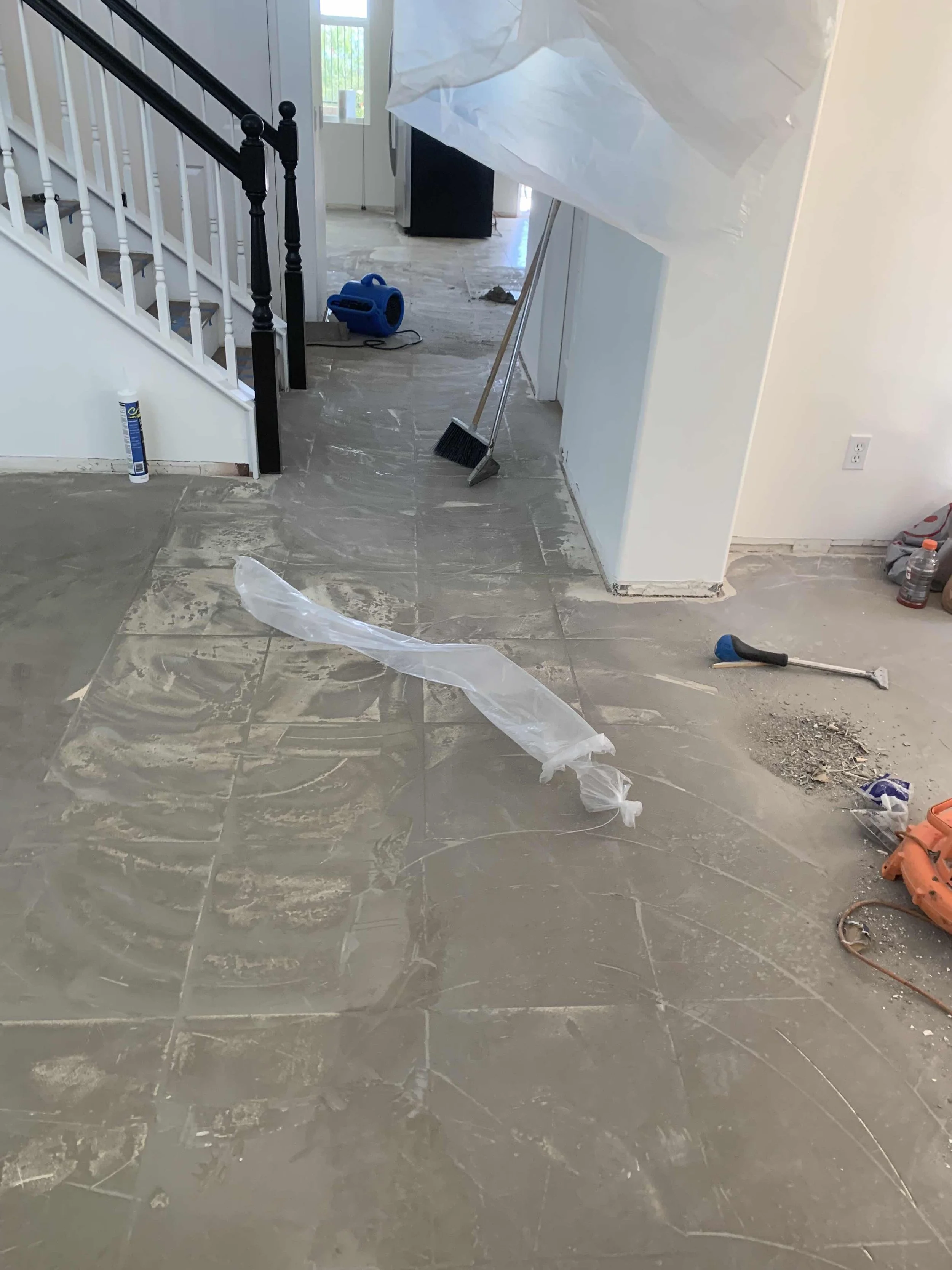 Construction site inside a house with tools and debris on the floor, including a broom, dustpan, plastic sheet, and bottles, indicating ongoing renovation or flooring work.