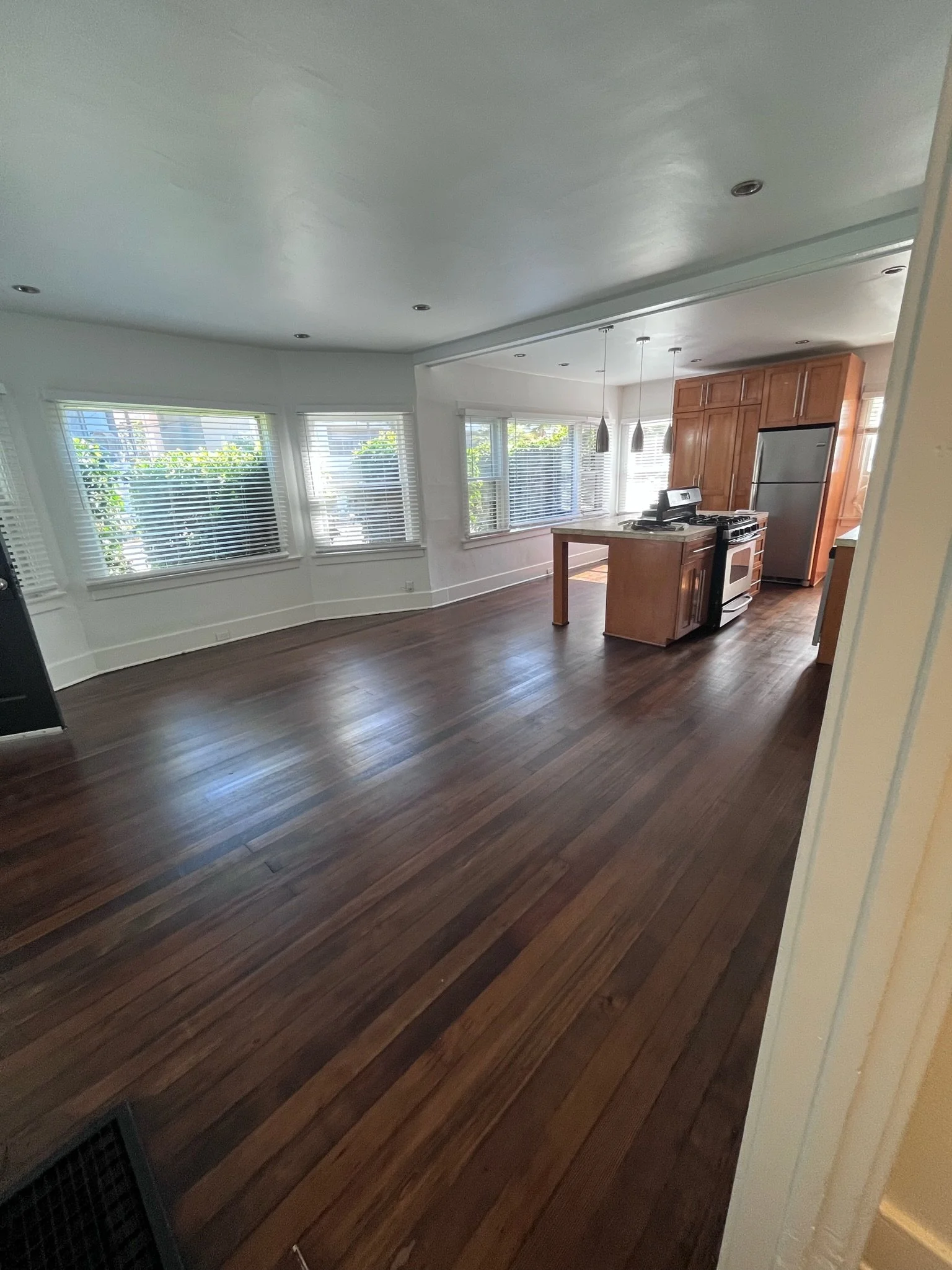 Empty living room with hardwood floors, large windows with blinds, and a kitchen area with wooden cabinets, stainless steel refrigerator, and a stove.