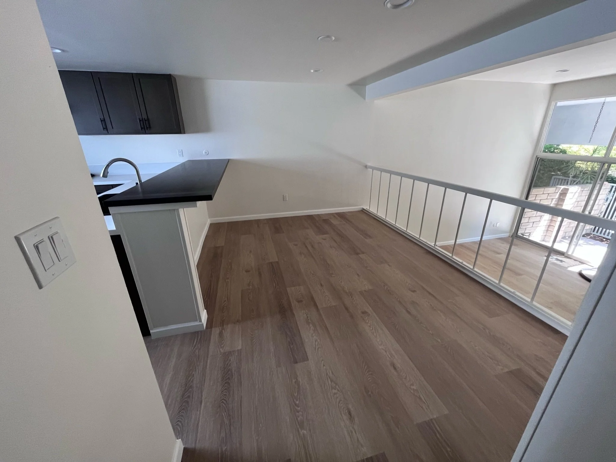 Empty living room with wooden floors, a glass sliding door, and kitchen area with dark cabinets and a black countertop