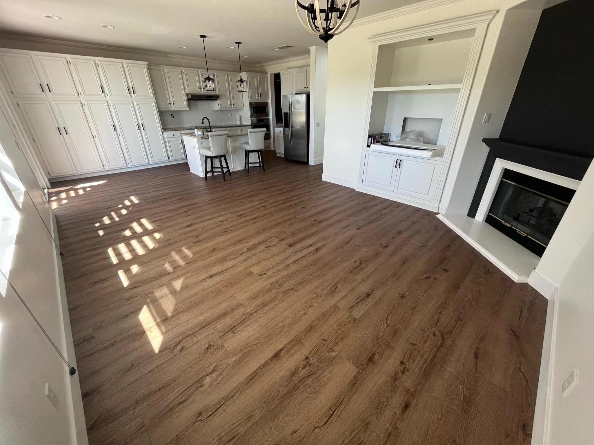 Empty living room with hardwood floors, white cabinetry in kitchen, and a black fireplace.