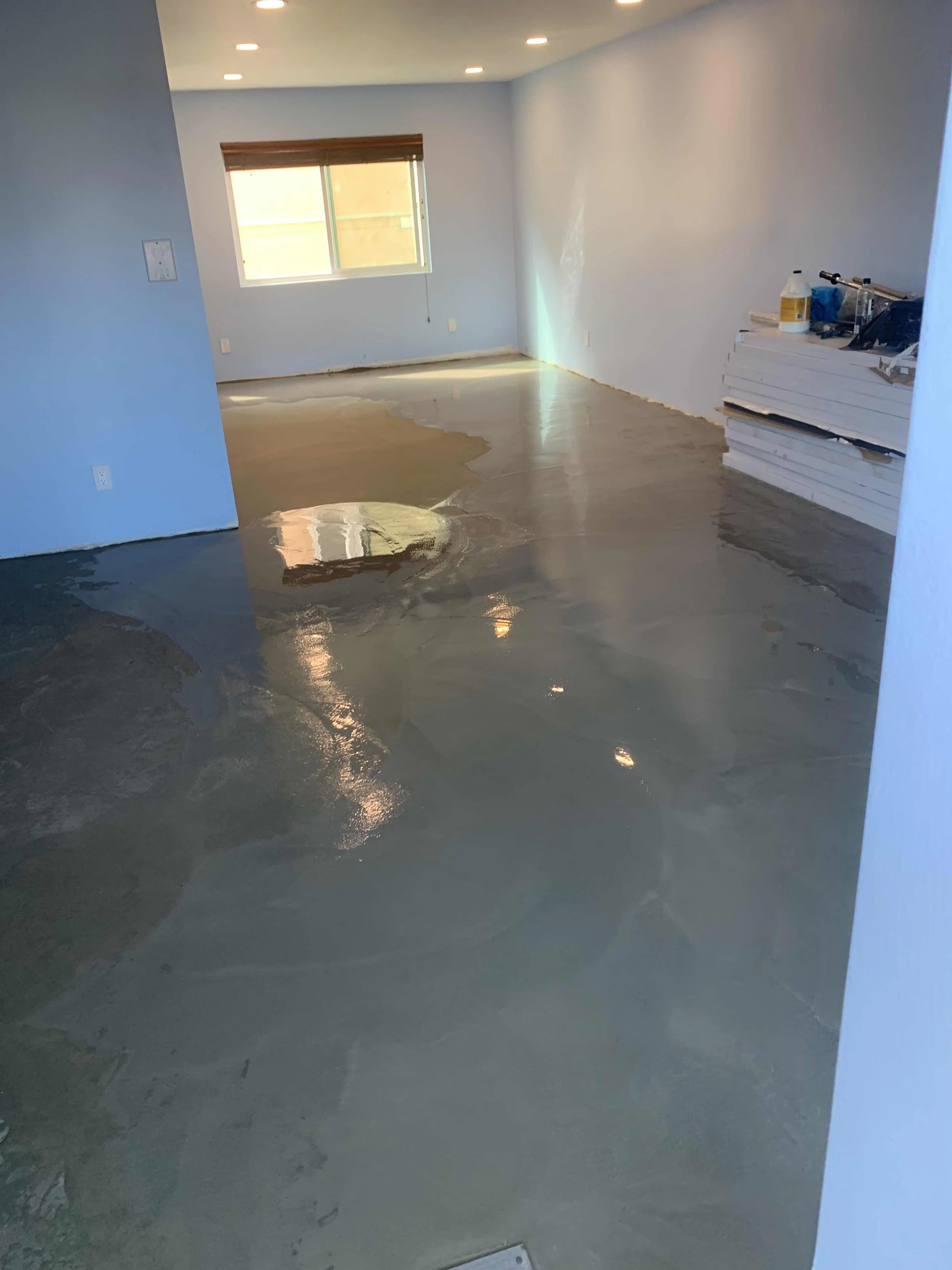 Empty room with new concrete flooring that is wet or polished, with a window on the far wall and construction materials on the right side.