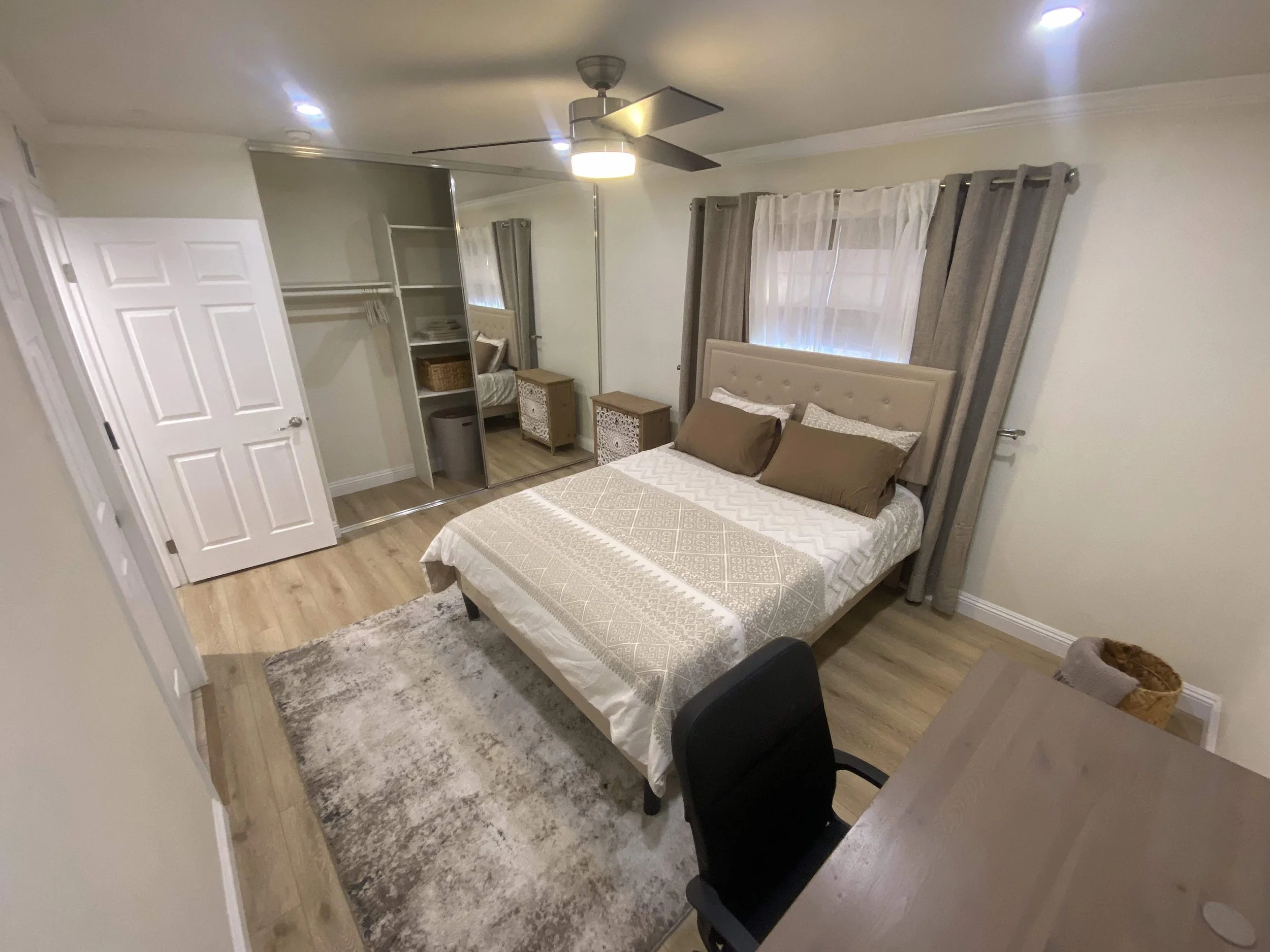 A bedroom with a bed, a mirror, nightstands, a closet with mirrored sliding doors, a ceiling fan, a window with curtains, a chair, a desk, and a basket.