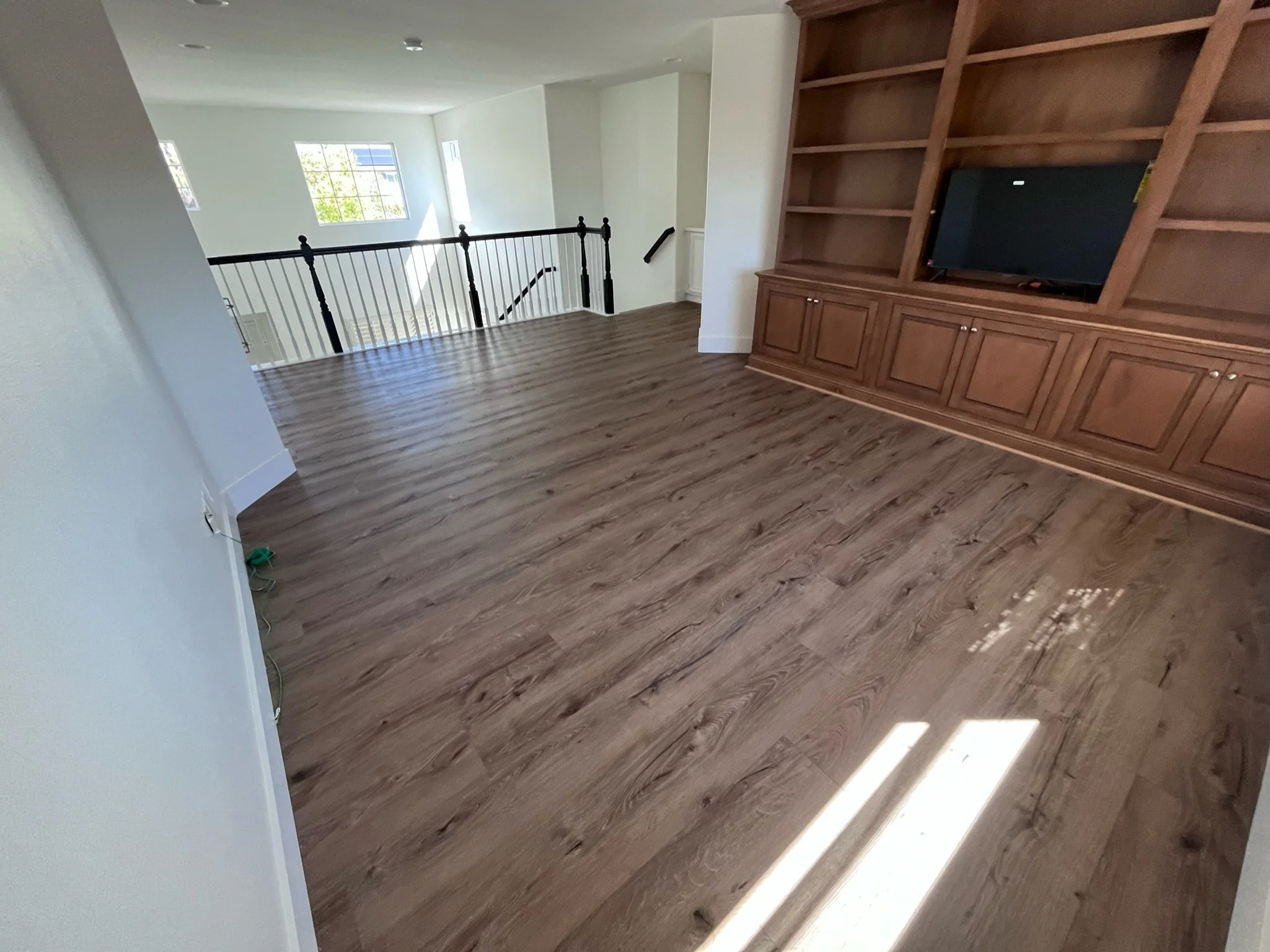 Empty room with hardwood floors, a built-in wooden wall unit with a TV, stairs with black railing, and large windows providing natural light.