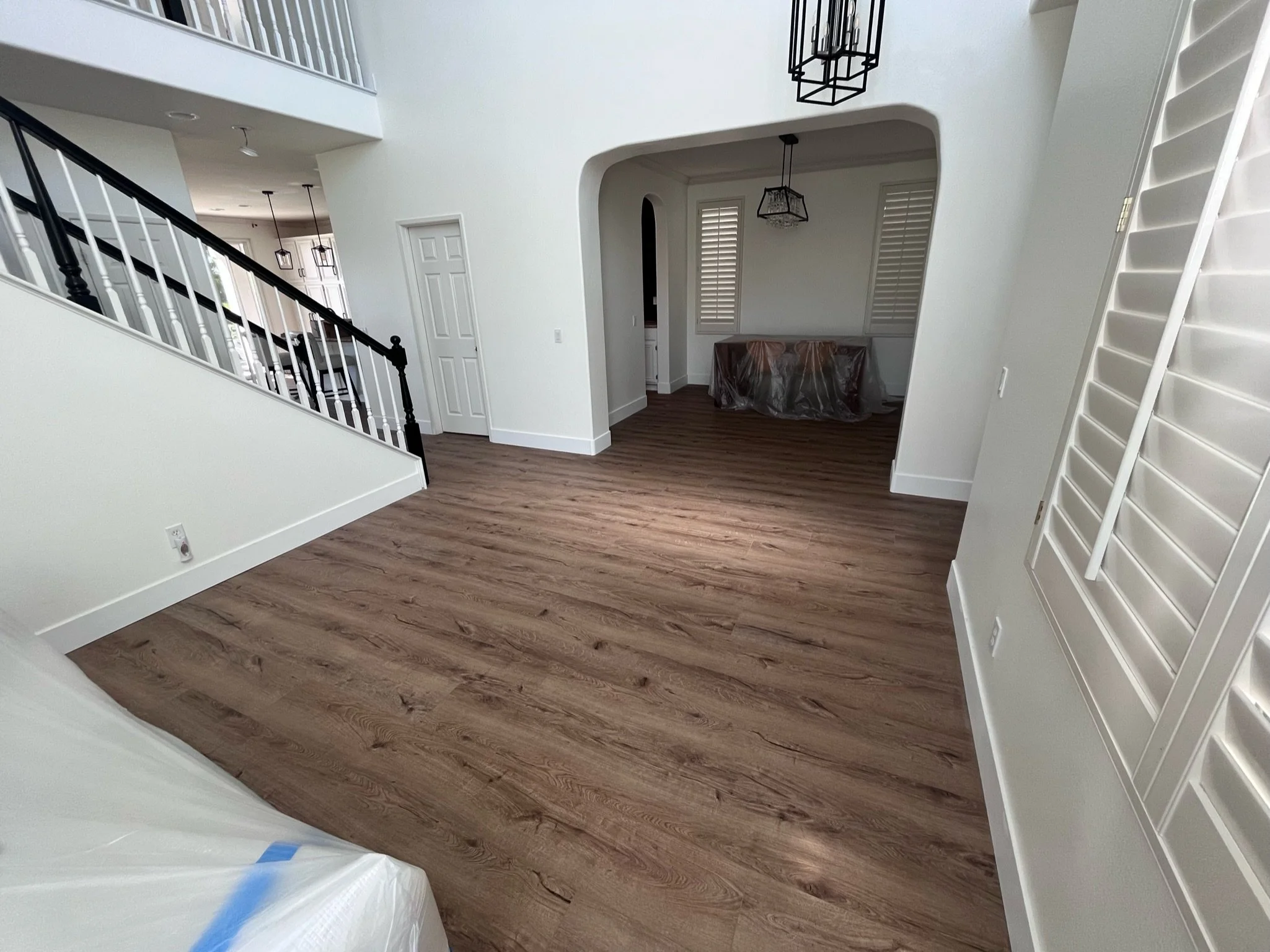 Empty living room with wood flooring, white walls, railing staircase on left, and a dining area with a covered table in the background.