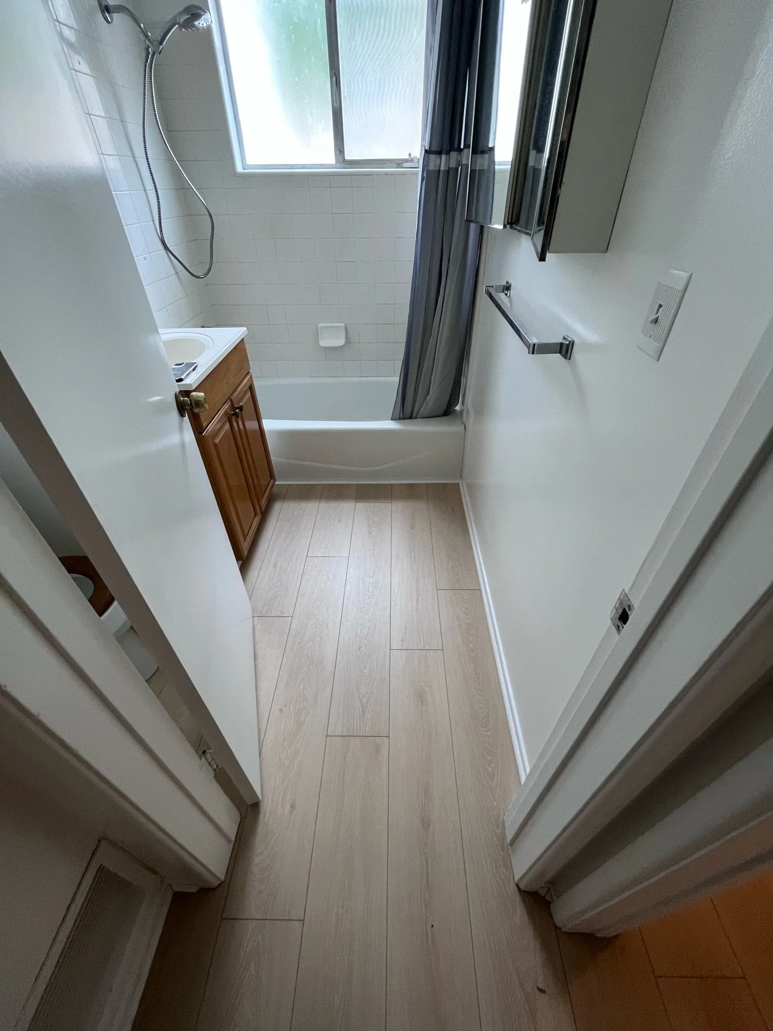 Small bathroom with a bathtub, a shower head, a window, a small wooden cabinet with a sink, a mirror cabinet, a towel rack, and a toilet partially visible on the left.