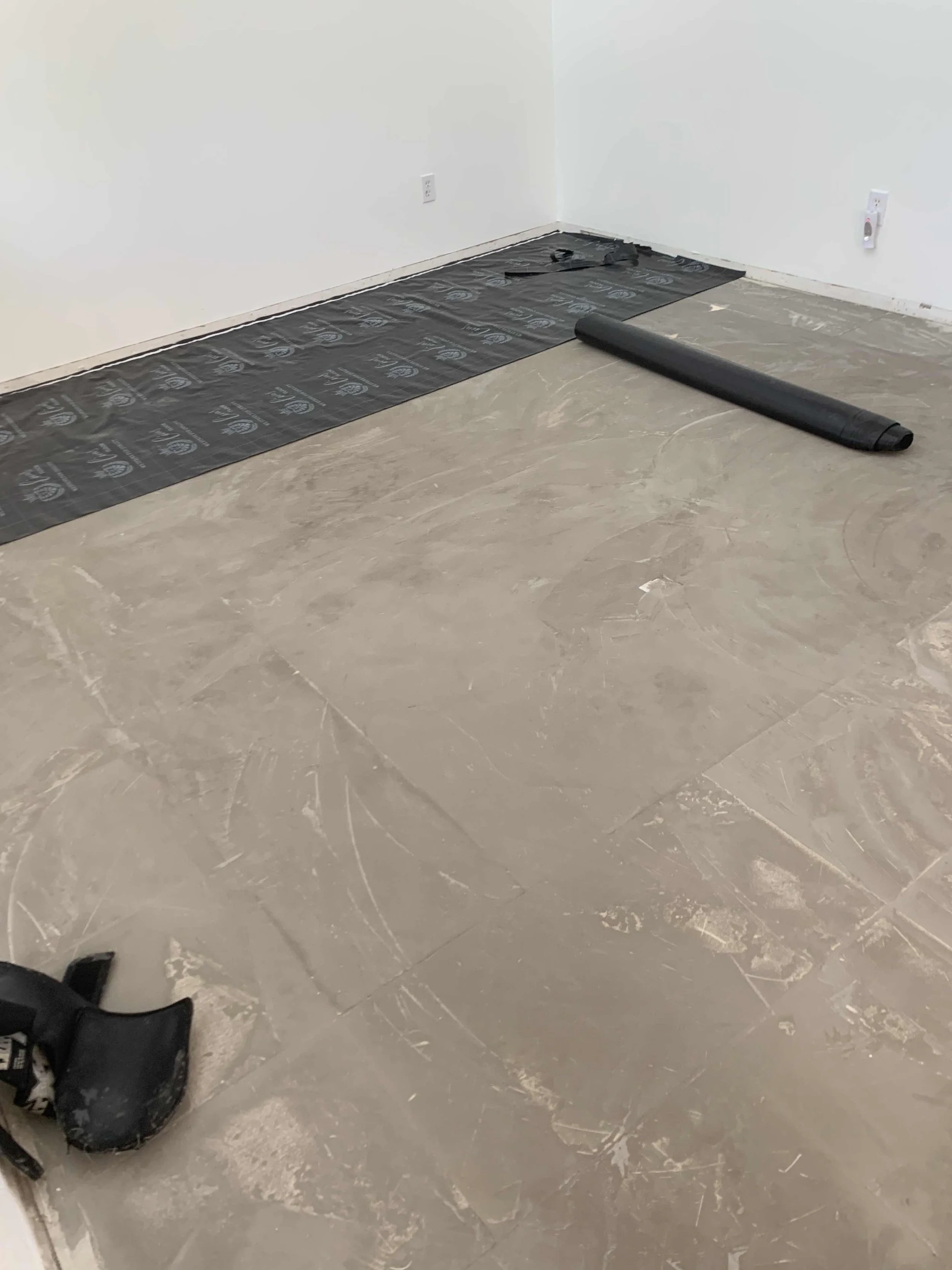 Empty room with a beige tiled floor, a black underlayment on the floor against the wall, and a rolled-up black carpet or padding.