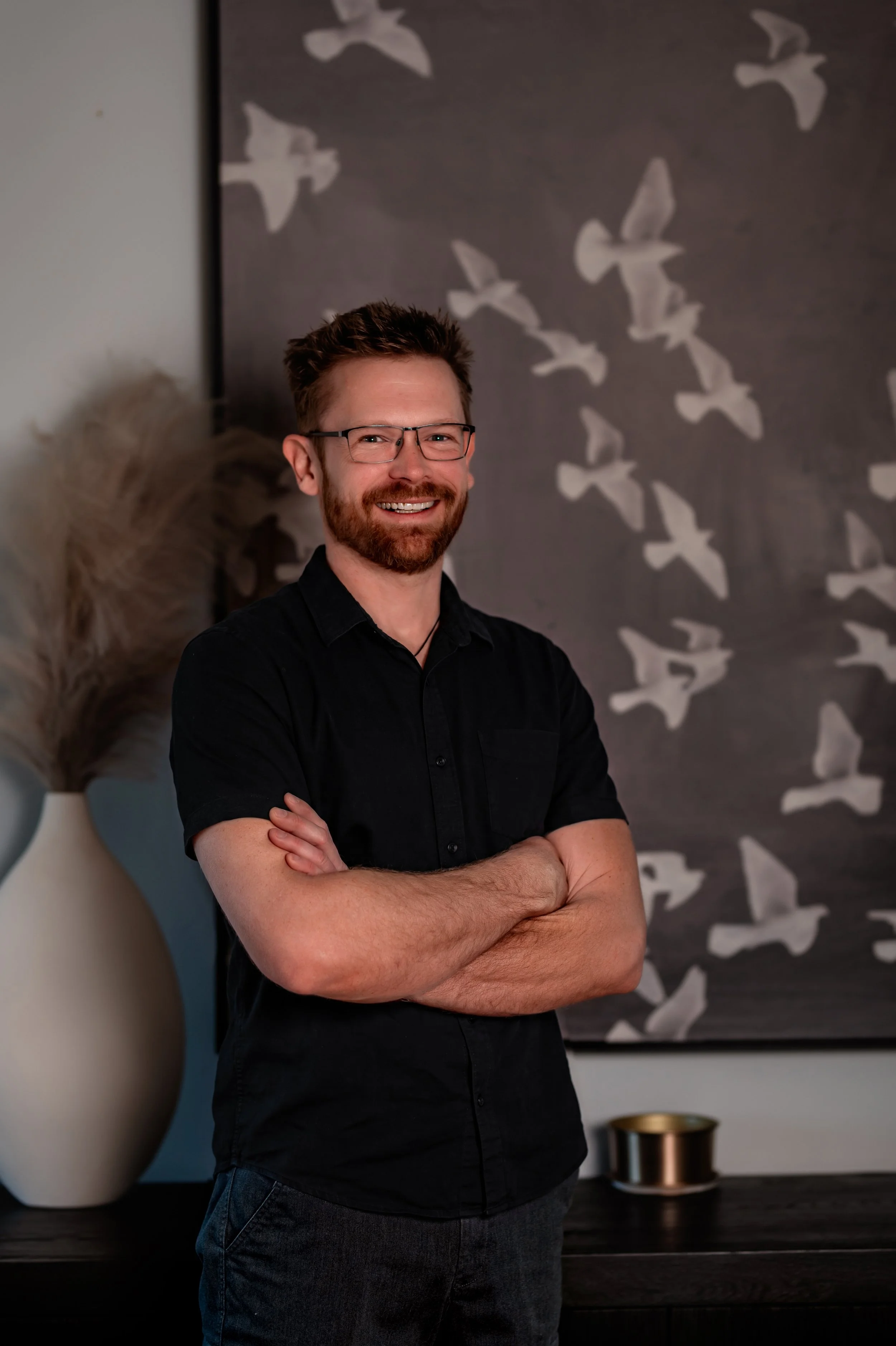 A man with reddish-brown hair, a beard, and mustache, wearing a dark long-sleeved shirt, standing with his arms crossed against a plain light gray background.