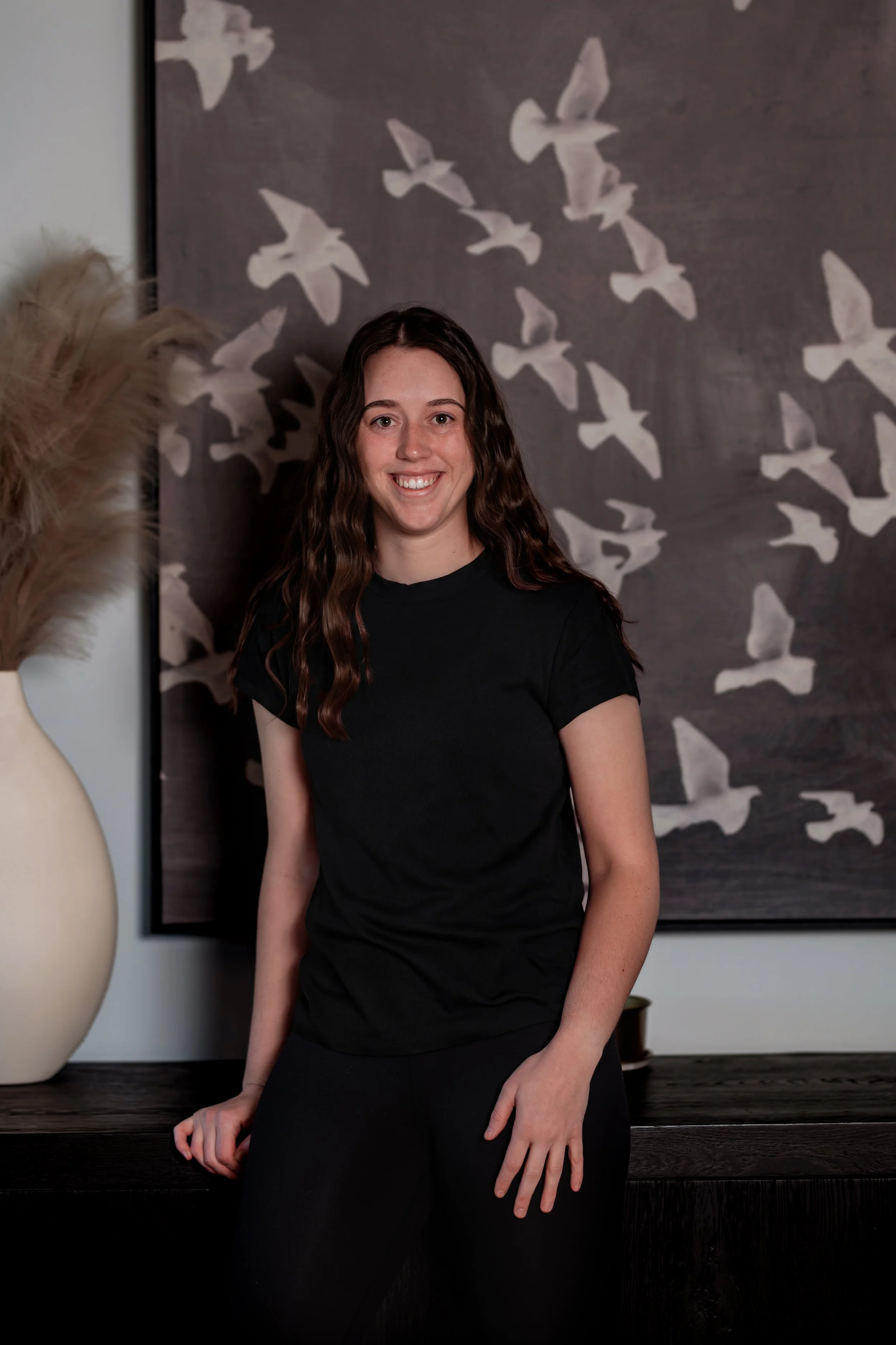 A woman with long brown hair, wearing sunglasses and a maroon hoodie, is sitting by a lake holding a fish she just caught. The background features a mountain, trees, and a partly cloudy sky.