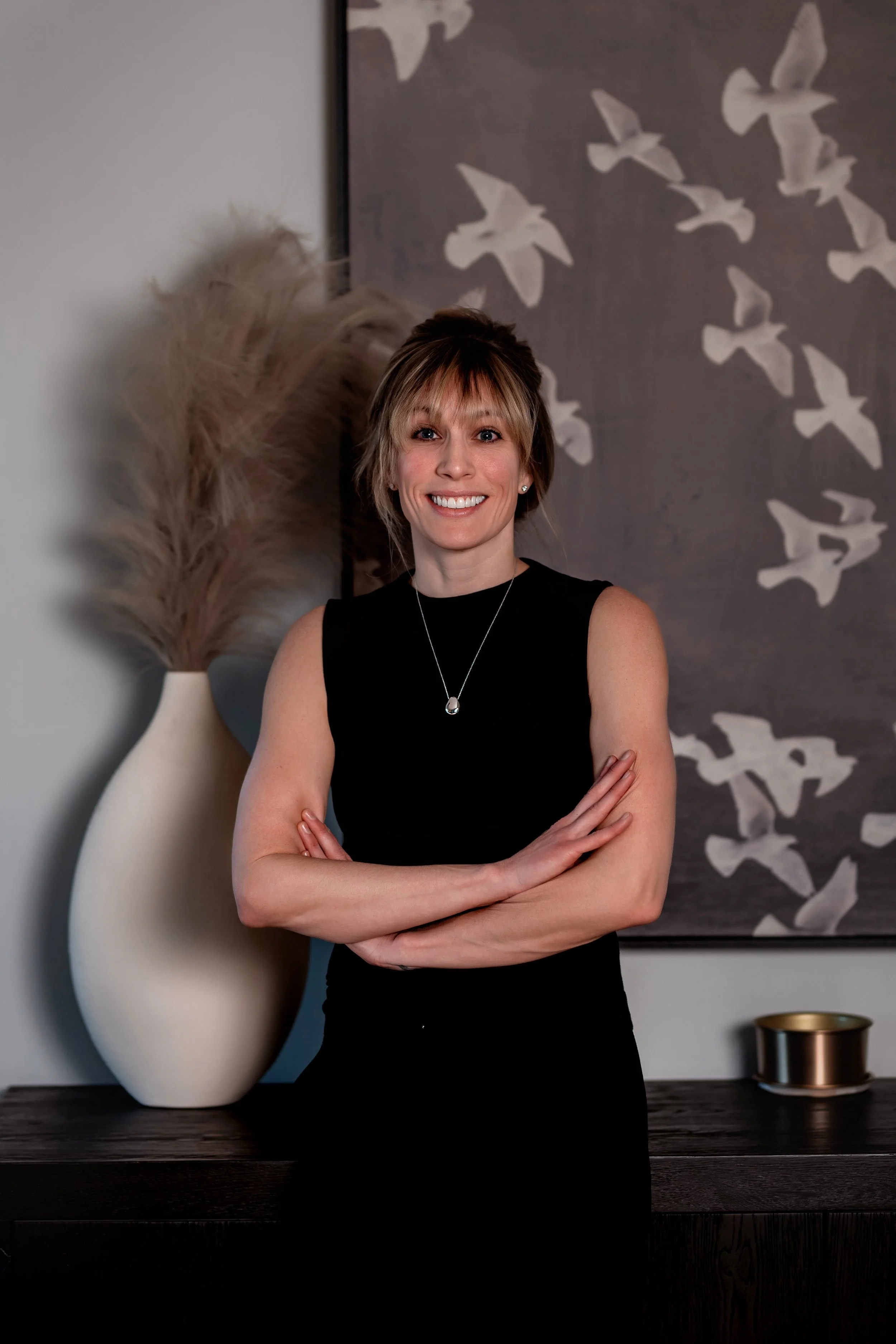 A woman with blonde, shoulder-length hair smiling, wearing a beige sleeveless top and blue jeans, standing indoors against a wall with white bird artwork and a large vase with pampas grass.