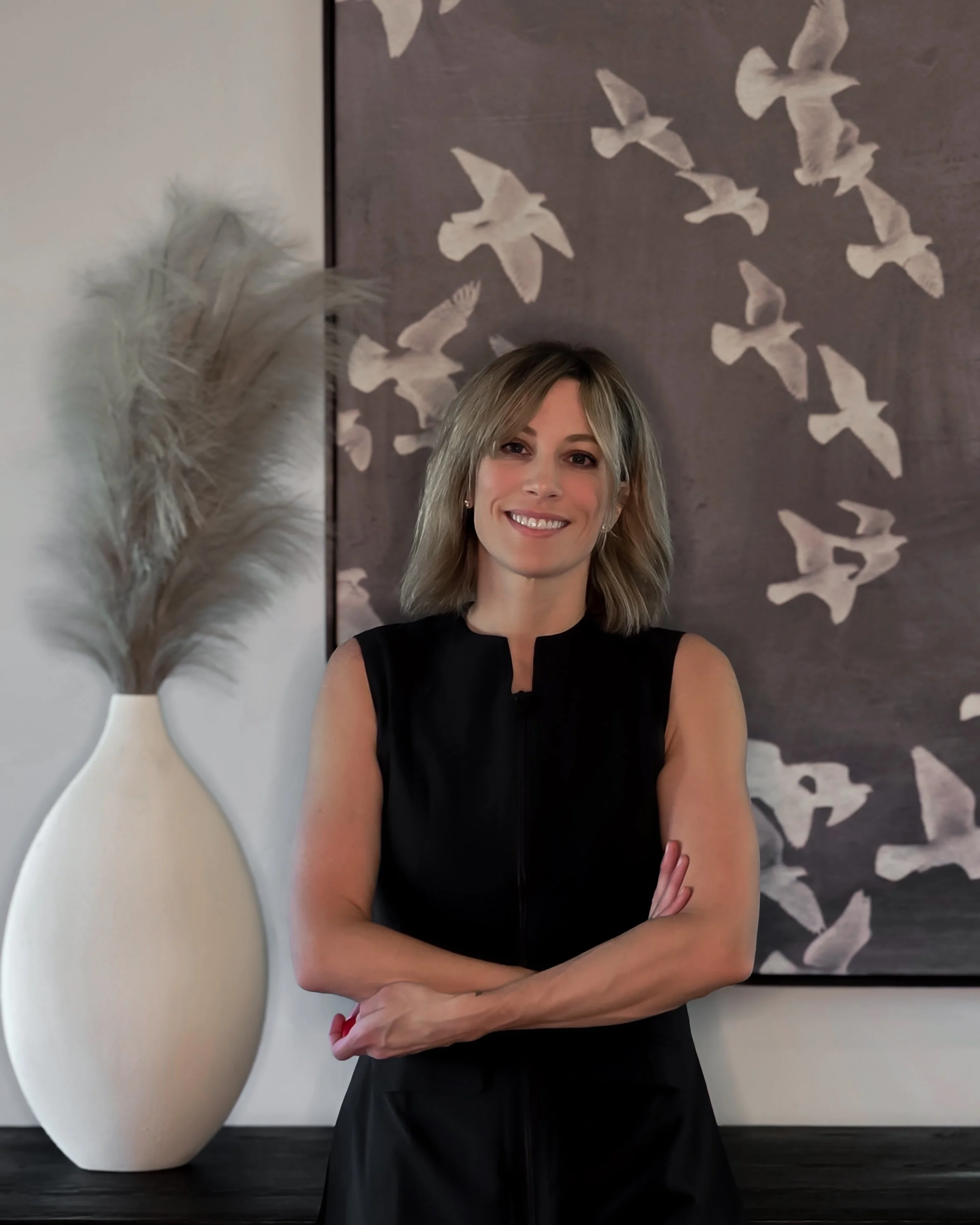 A woman with blonde, shoulder-length hair smiling, wearing a beige sleeveless top and blue jeans, standing indoors against a wall with white bird artwork and a large vase with pampas grass.
