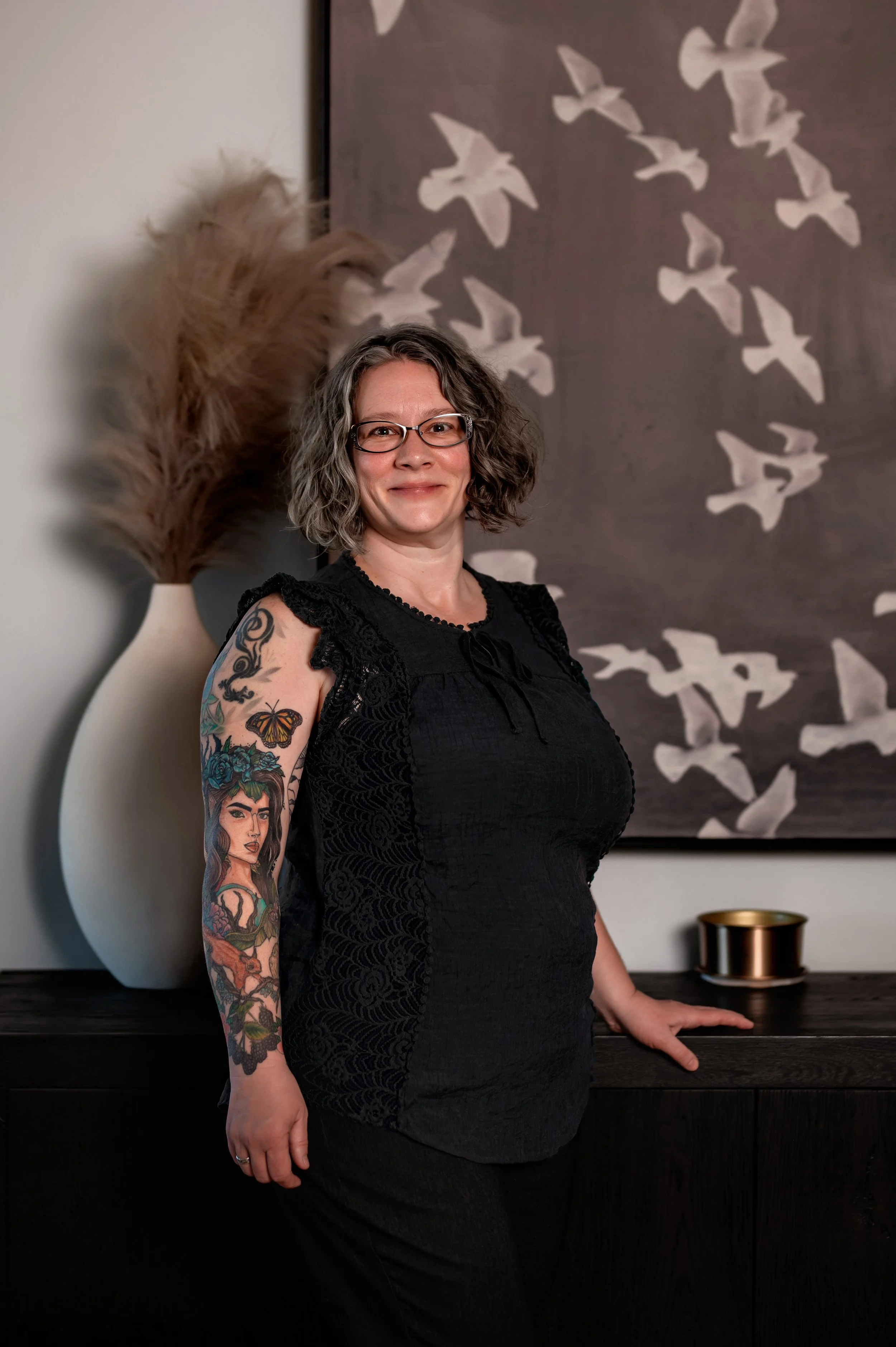 A woman with curly gray hair and glasses smiling outdoors in a forest setting.
