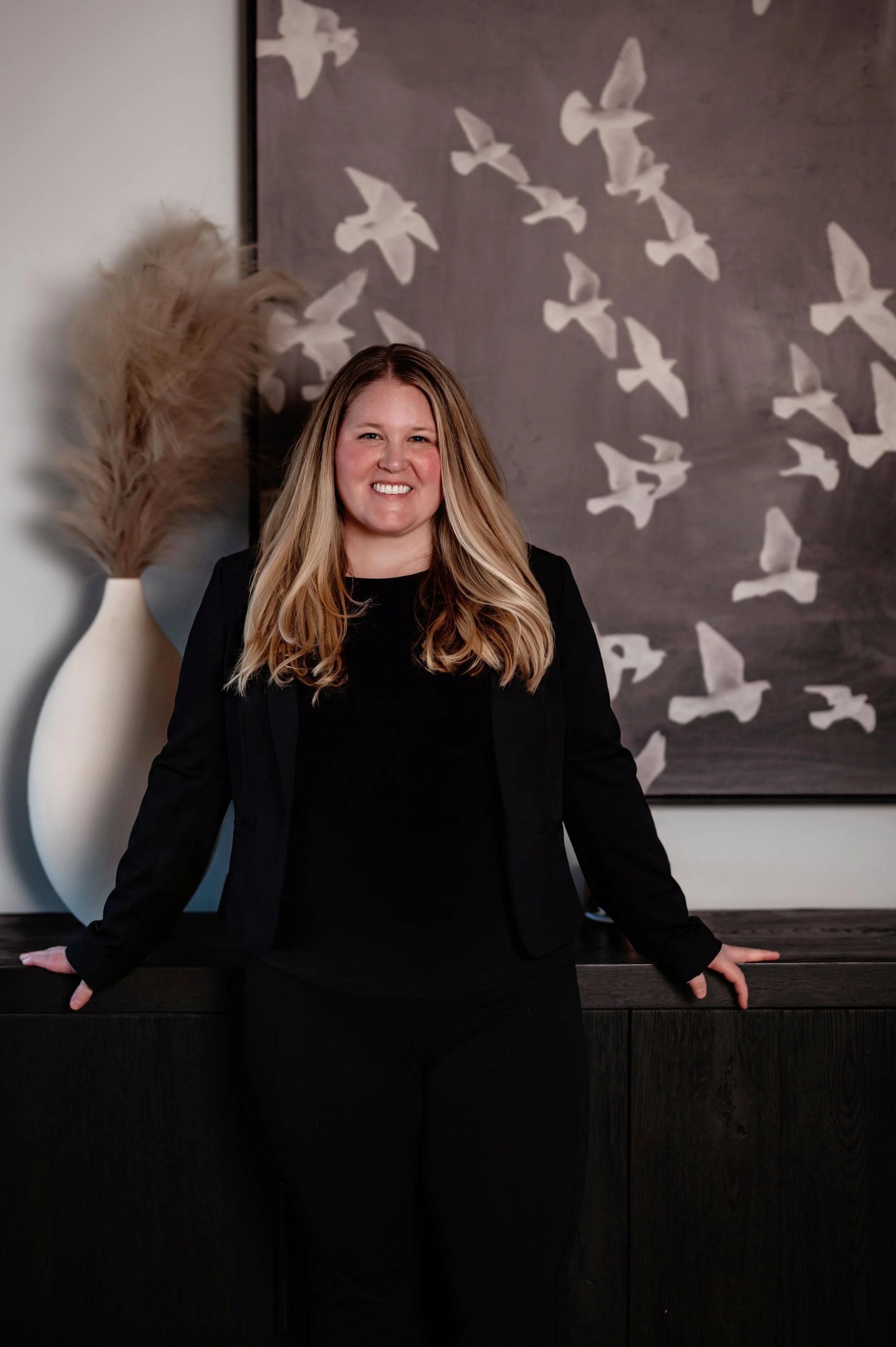Portrait of a woman with long wavy hair smiling at the camera, standing against a plain wall.