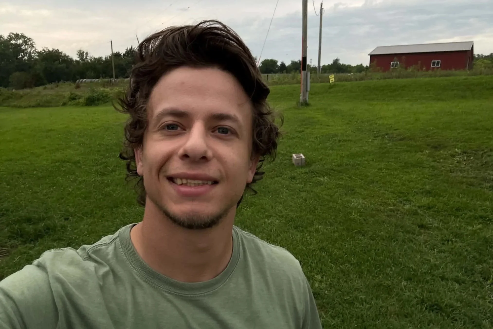 A young man taking a selfie outdoors in a grassy area with a red barn and utility poles in the background.