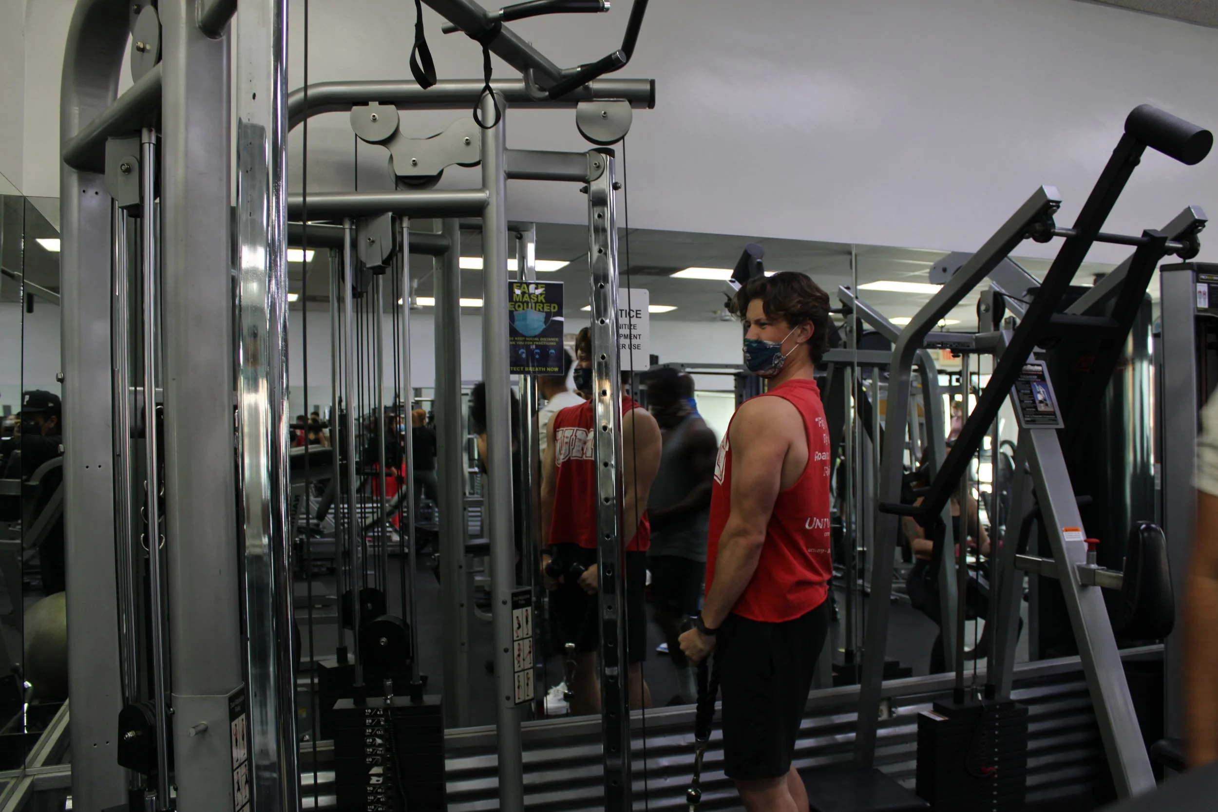 A man wearing a red tank top and black shorts standing in a gym, wearing a face mask, holding ropes used for strength training, with gym equipment and other people working out in the background.