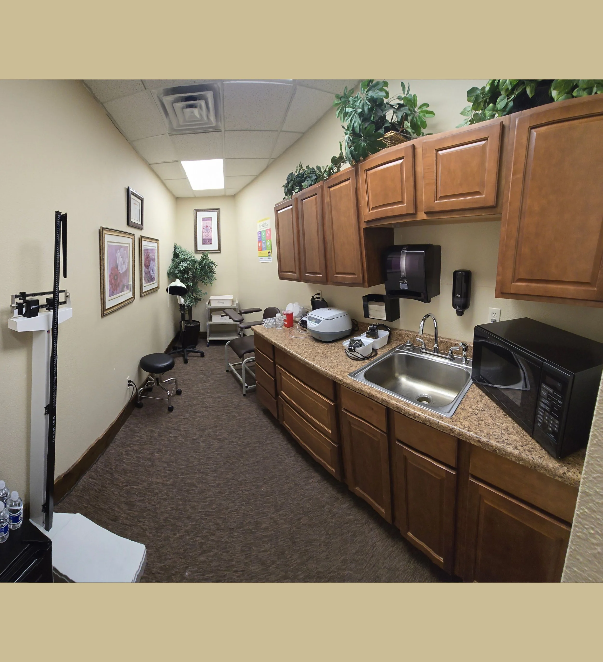 Medical office treatment room with wooden cabinets, a small sink, microwave, and various medical and office equipment on the countertop; framed artwork on the wall; chairs and a scale in the background.