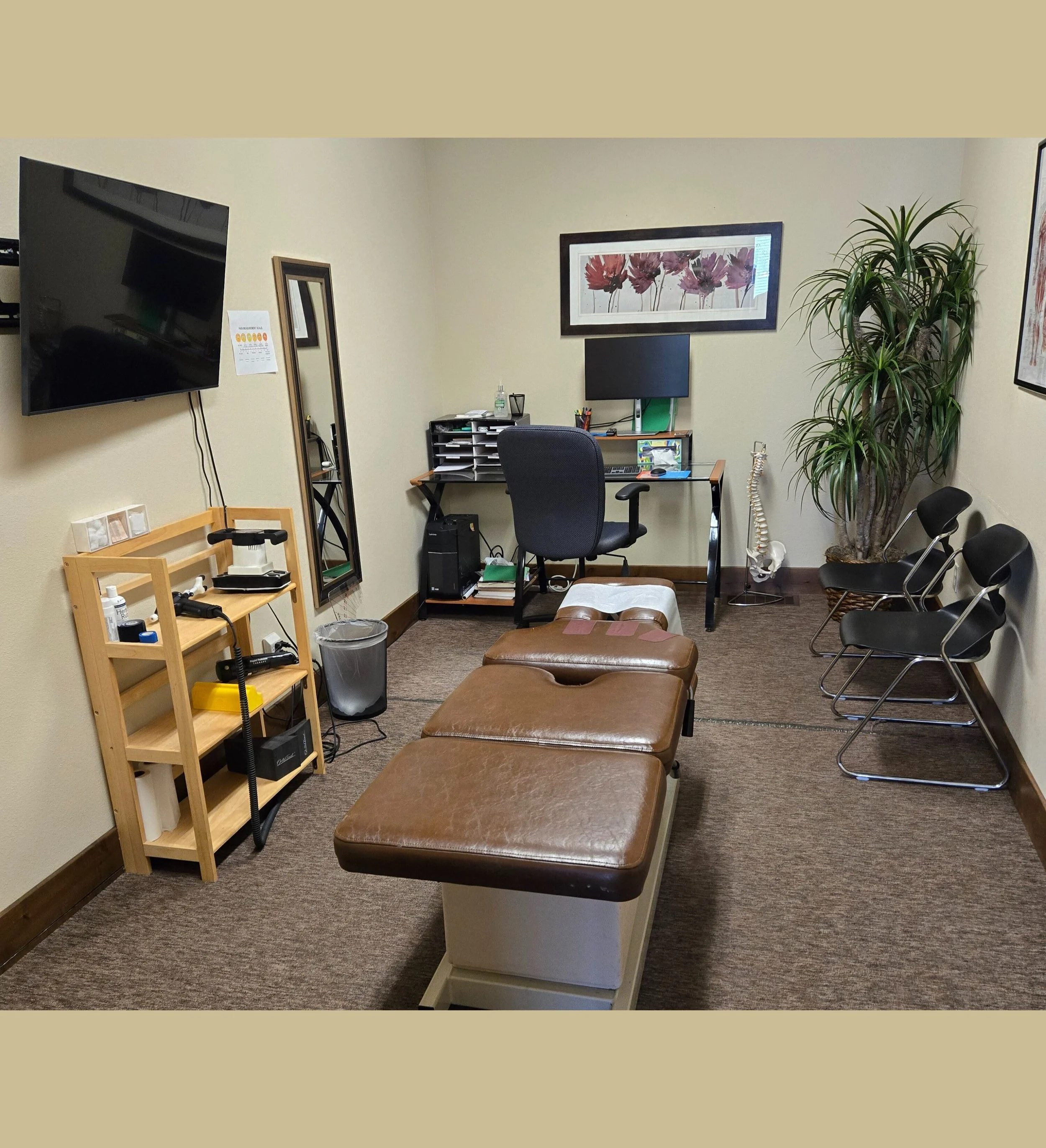 Medical examination room with a massage table, a desk with a computer, chairs, a wall-mounted TV, and various medical equipment and decor.