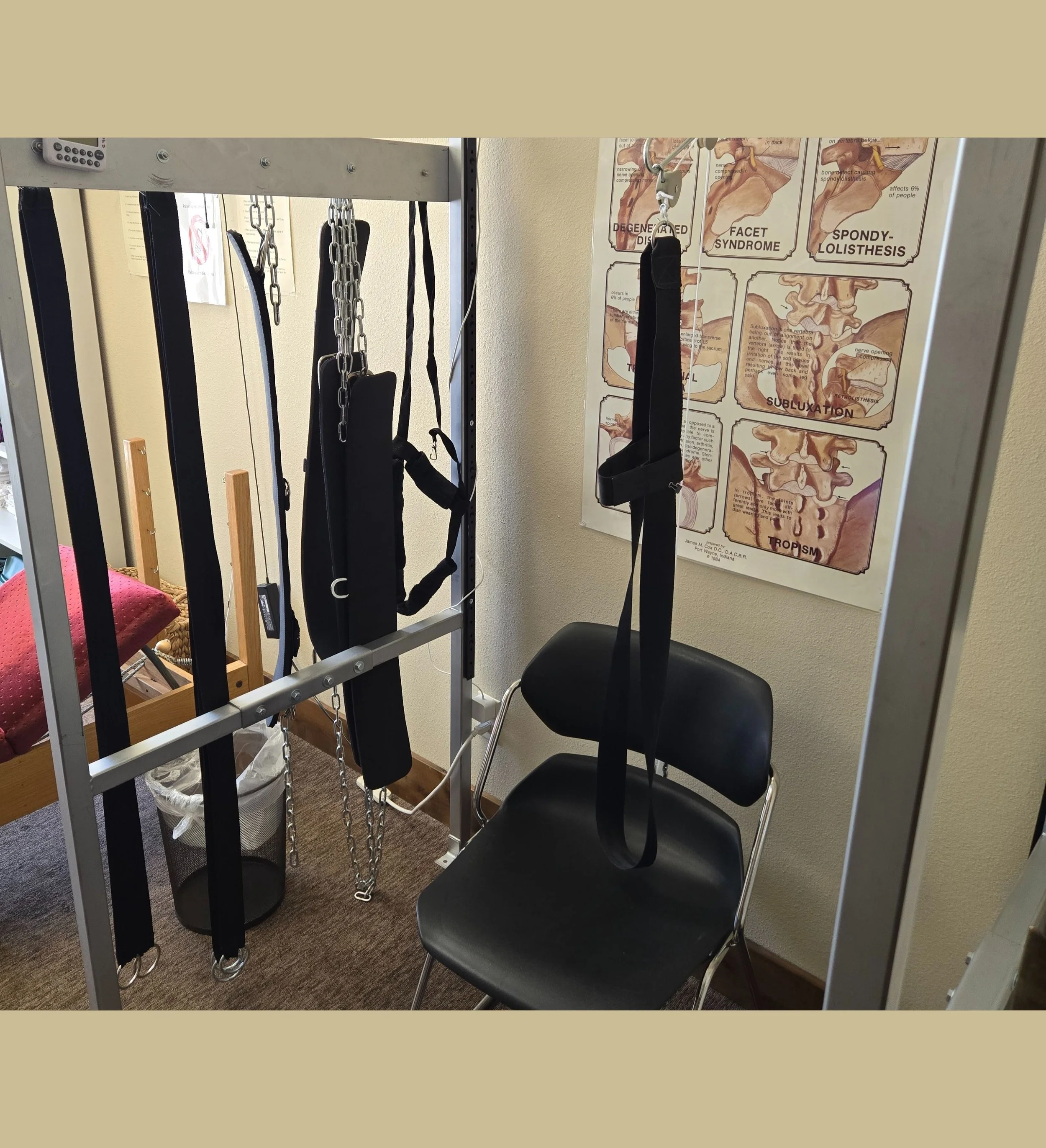 Physical therapy room with suspension straps and harnesses, medical poster on spinal issues, black chair, and a trash can.