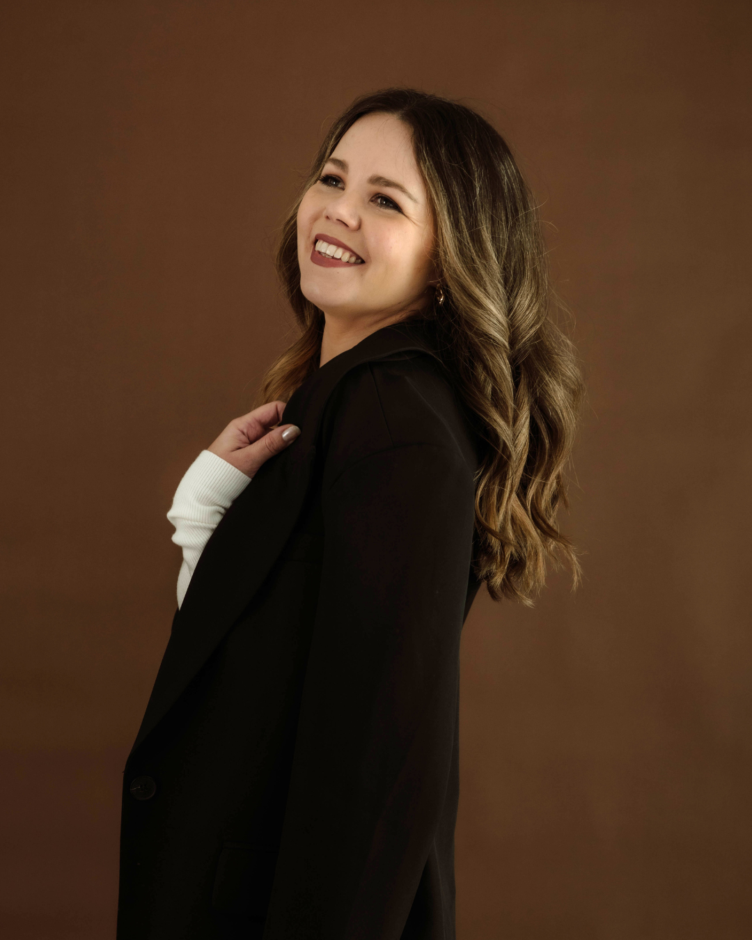 Smiling woman with wavy hair wearing a black blazer and white shirt, posing against a brown background.