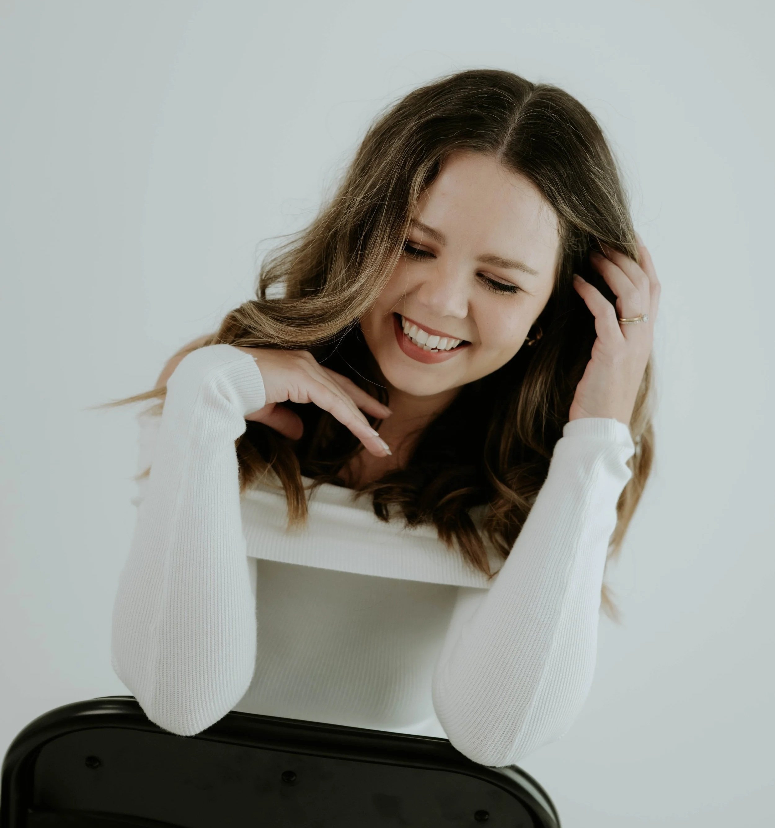 A woman with brown, wavy hair and a bright smile, wearing a white long-sleeve shirt, touching her hair with one hand, against a plain white background.
