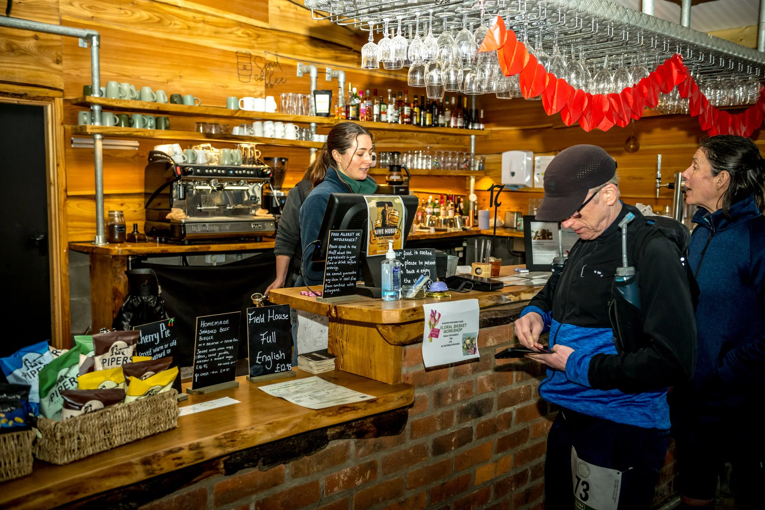 People at a cafe counter with a coffee machine and menu board in view, wooden shelves, hanging glasses, and snacks on display.
