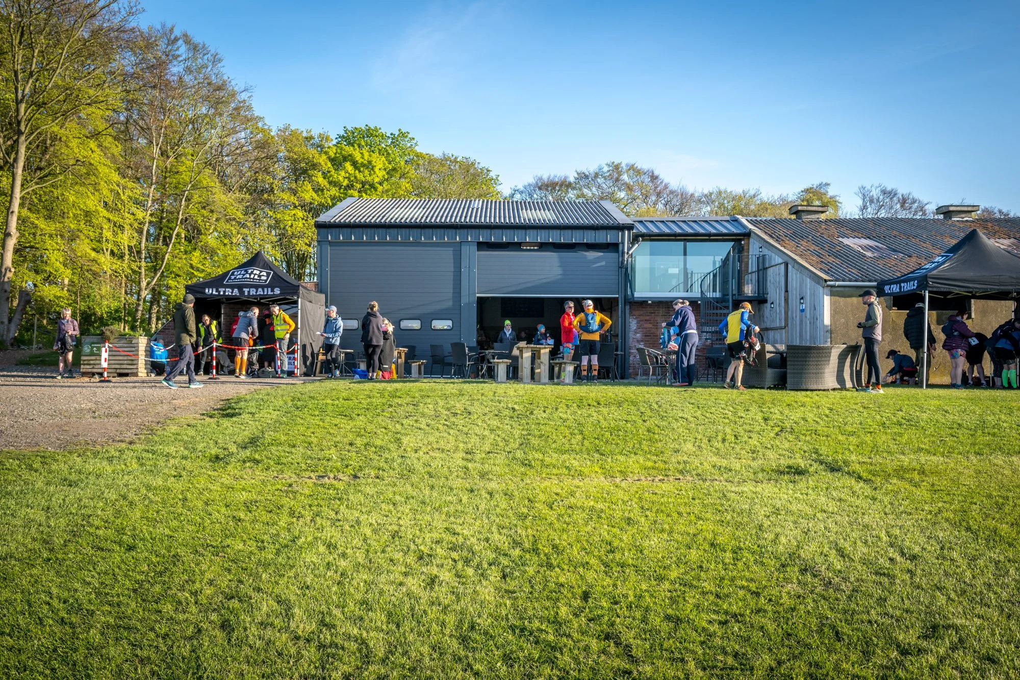 People gathered outside a building with two tents marked 'Ultra Trails,' some standing, sitting, or preparing bicycles on a grassy field in a wooded area on a sunny day.