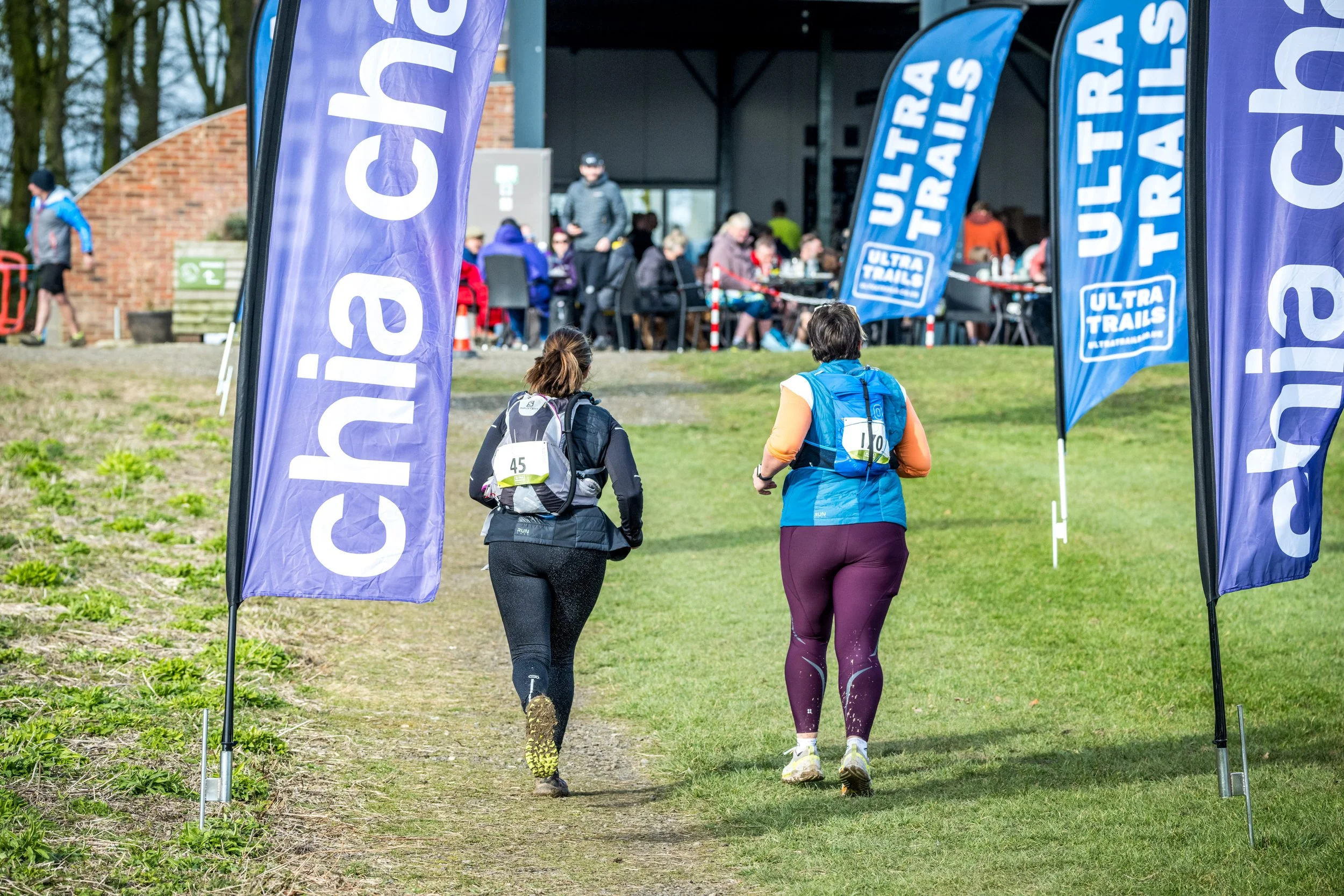 Two participants with numbered bibs running in an outdoor event with 'chia charge' and 'ultra trails' banners visible.