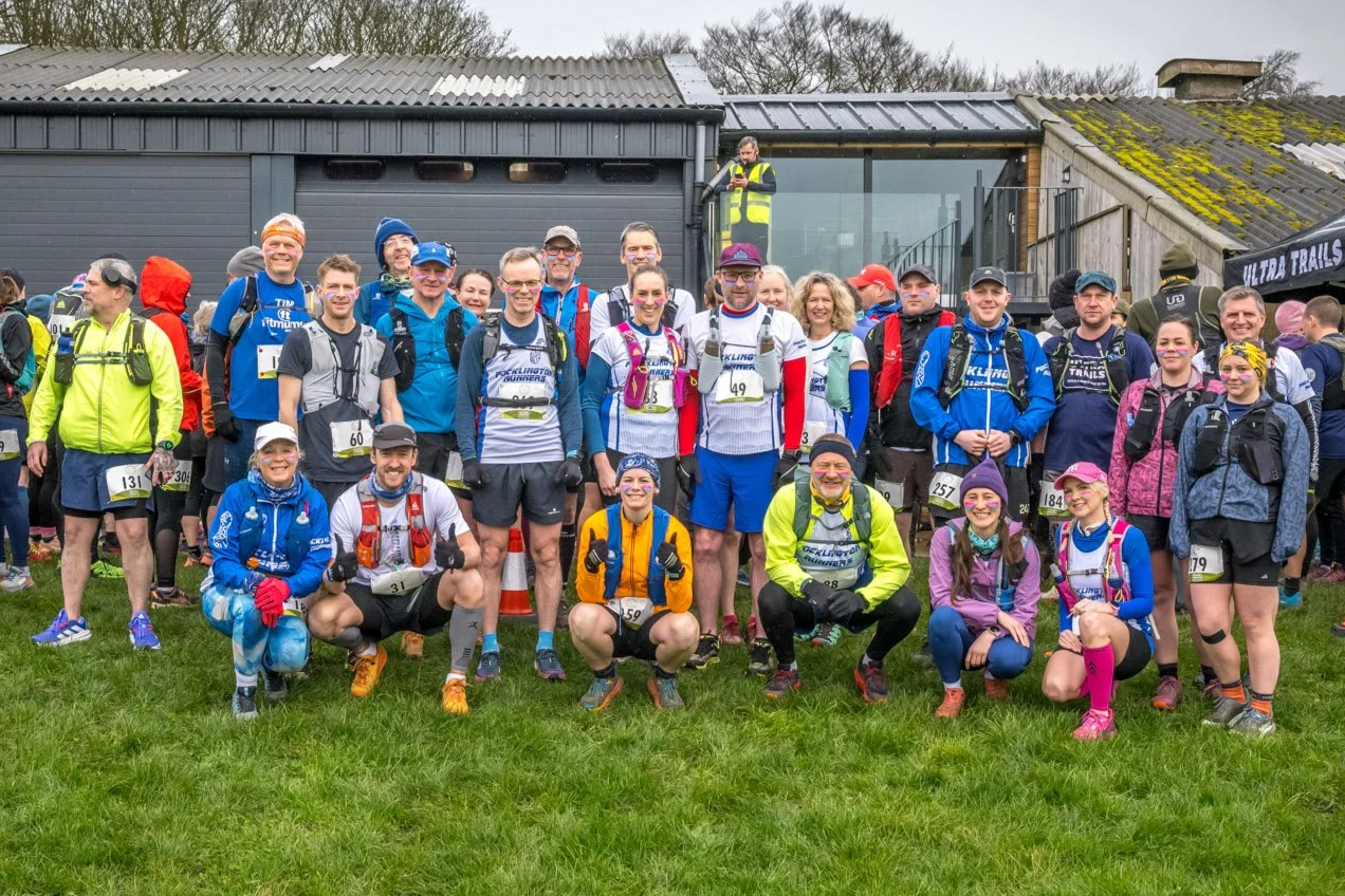 Group of runners gathered outdoors for a race event, many wearing athletic gear and numbered bibs, with some smiling and giving thumbs up.