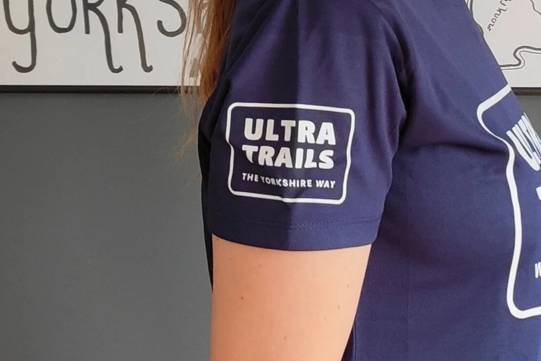 Person wearing a navy blue shirt with "Ultra Trails The Yorkshire Way" logo on sleeve; partial view of wall decor in background.