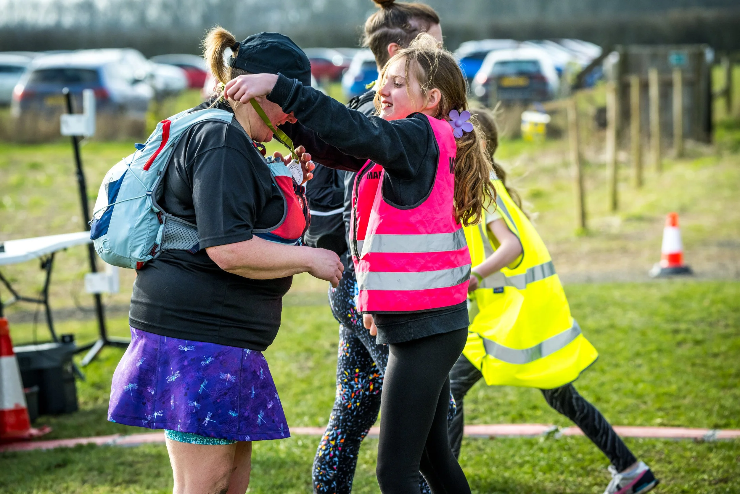A young girl in a pink reflective vest places a medal around the neck of an adult woman wearing athletic gear, a blue backpack, and a purple skirt. The setting appears to be an outdoor event or race, with parked cars and other participants in the bac