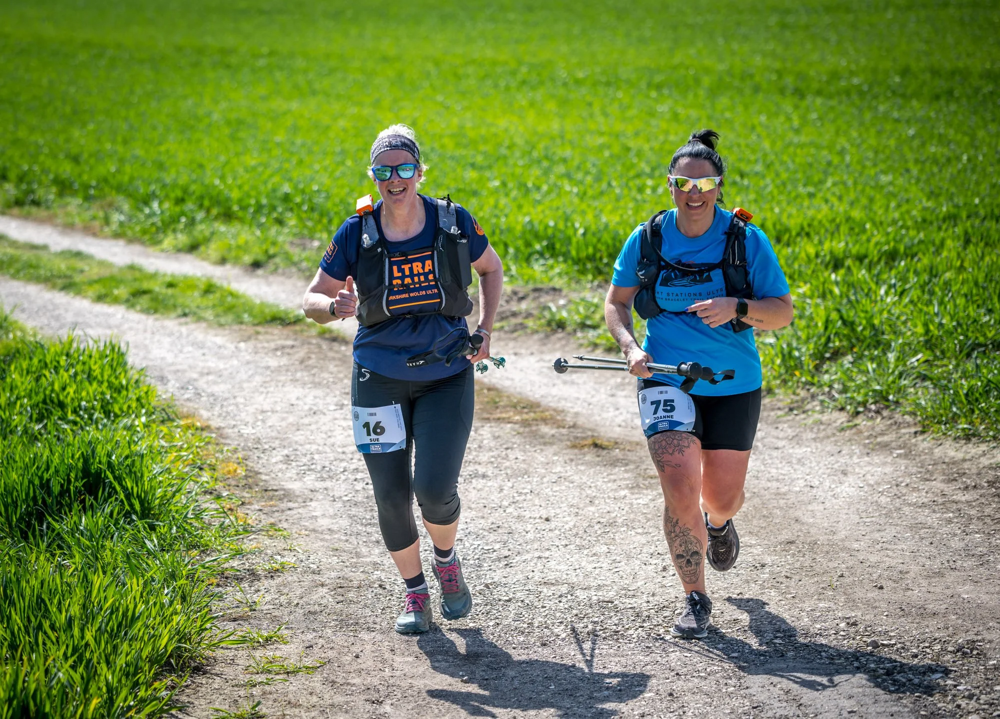 Two women running on a dirt trail through a green field, wearing athletic clothes and sunglasses, with race bibs numbered 16 and 75.