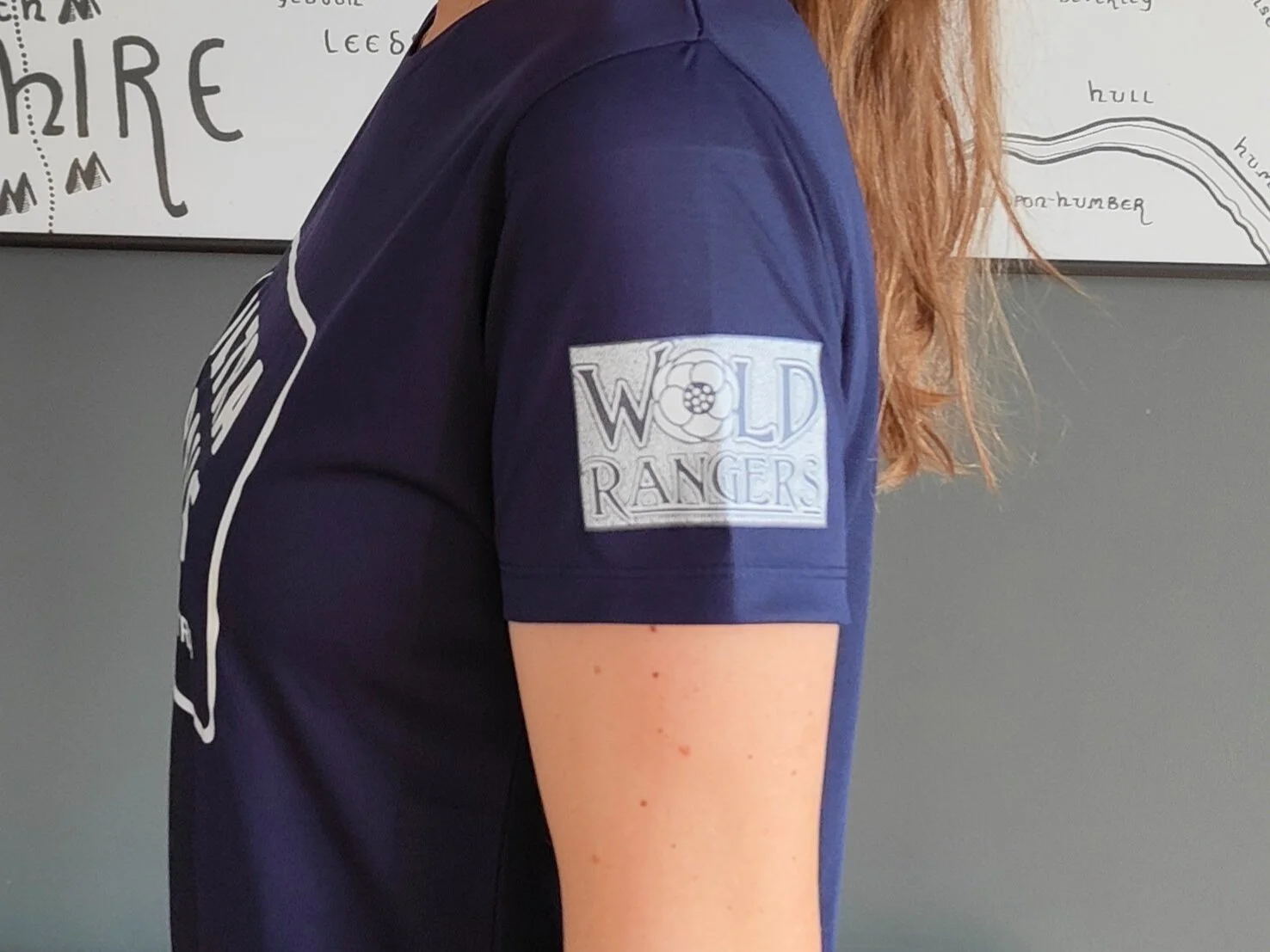 Person wearing a navy shirt with "Wold Rangers" patch on the sleeve, standing in front of a map.