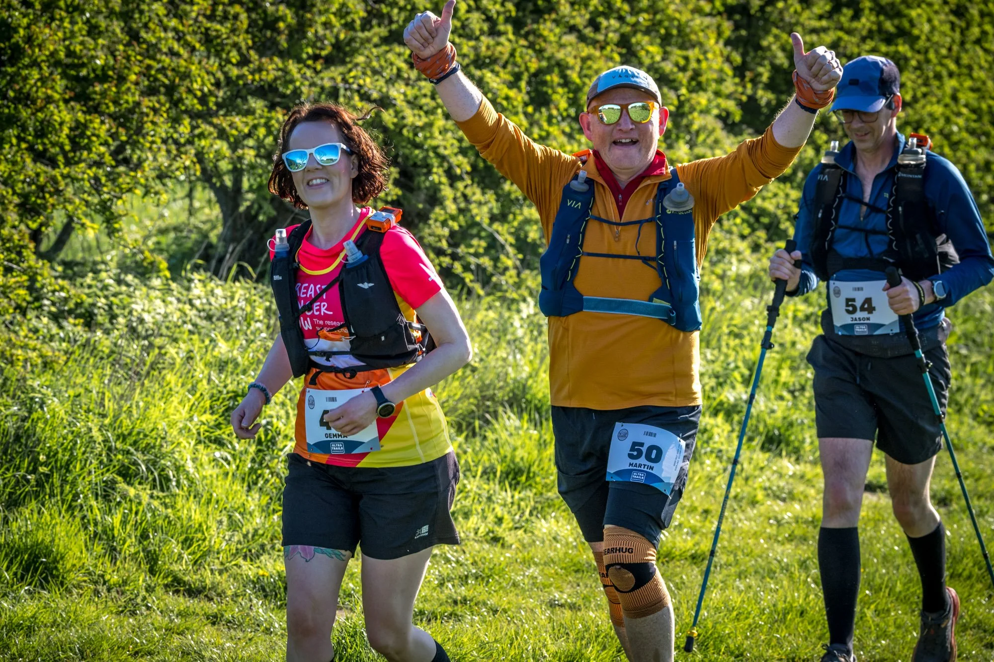 Group of three people participating in a trail race in a green outdoor setting, wearing athletic gear and numbered bibs, with the middle person making a thumbs-up gesture.
