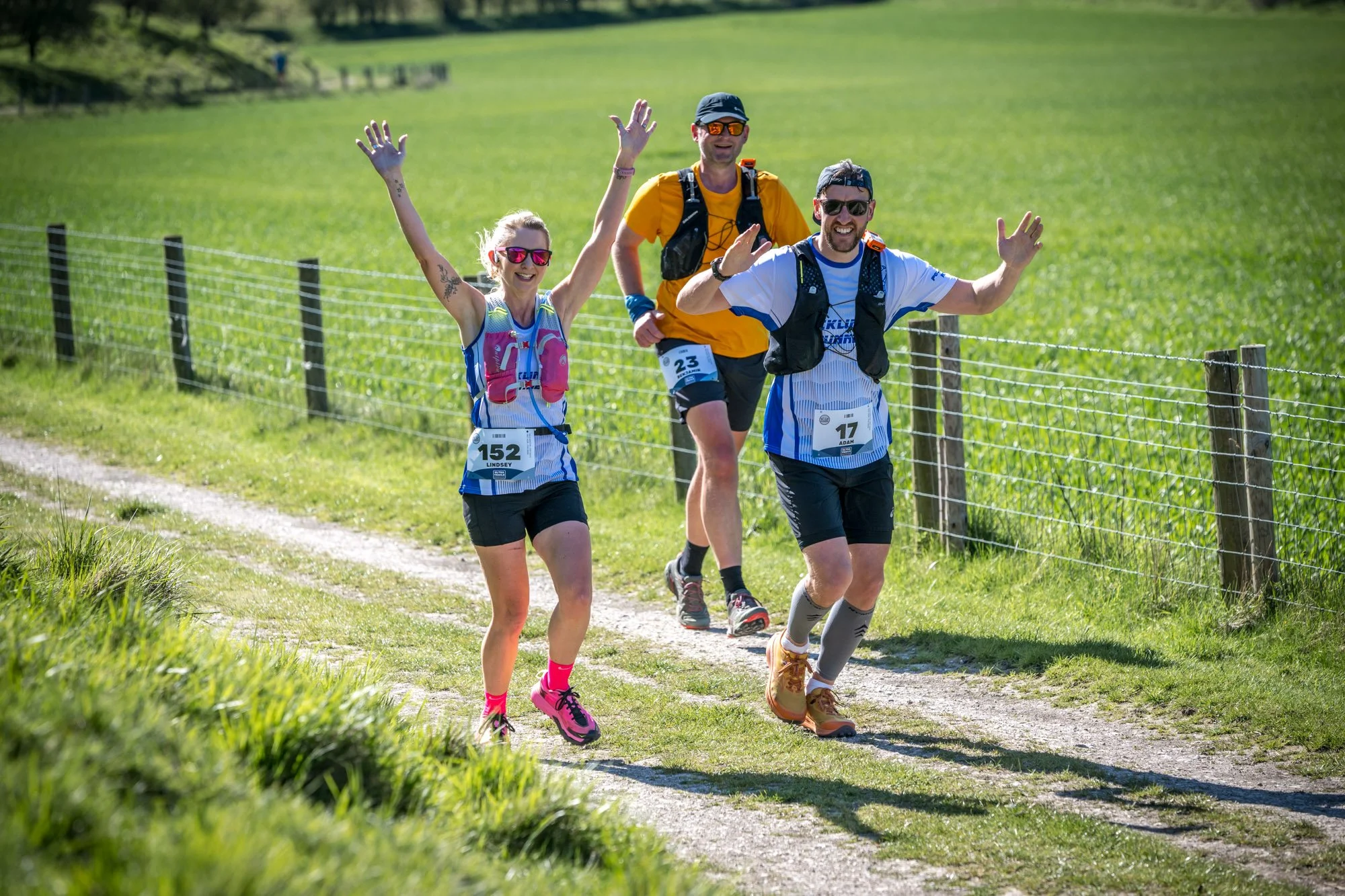 Three people running on a dirt trail in a rural, green landscape, smiling and waving at the camera, dressed in athletic clothing and wearing race bibs.