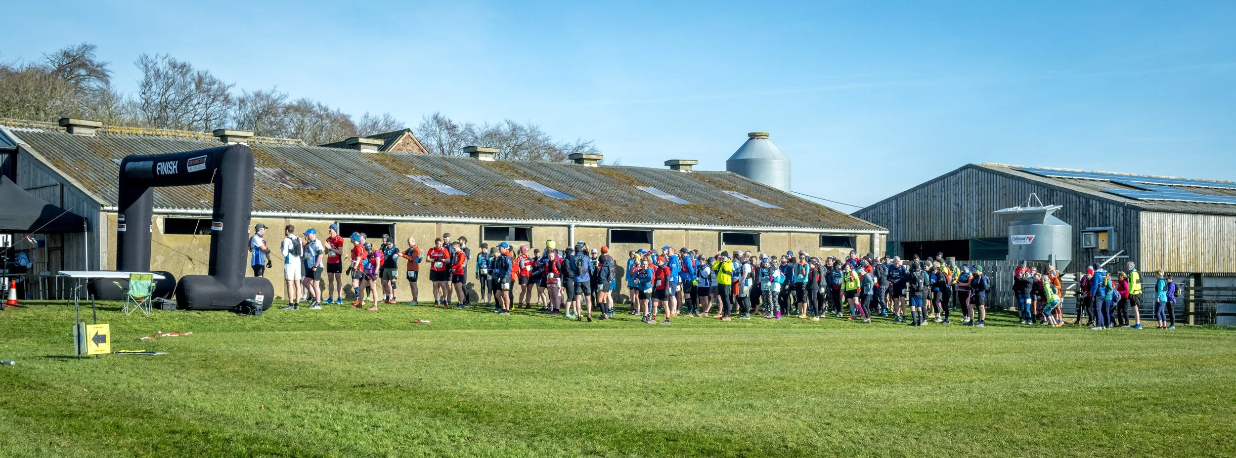 Group of runners gathered at a finish line next to a farm building, with clear blue sky in the background.