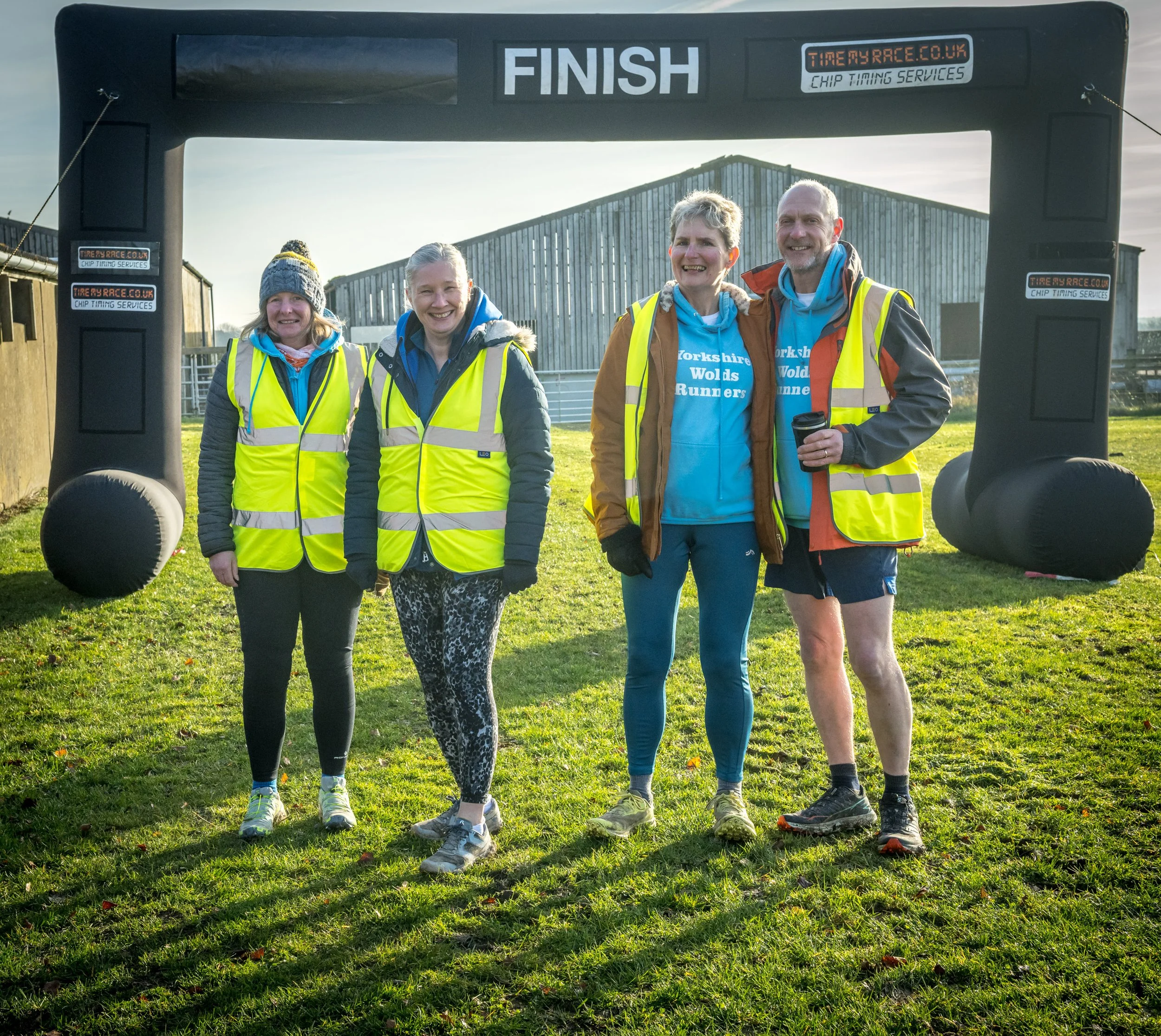 Four people in reflective vests standing in front of a race finish line banner in an outdoor setting.