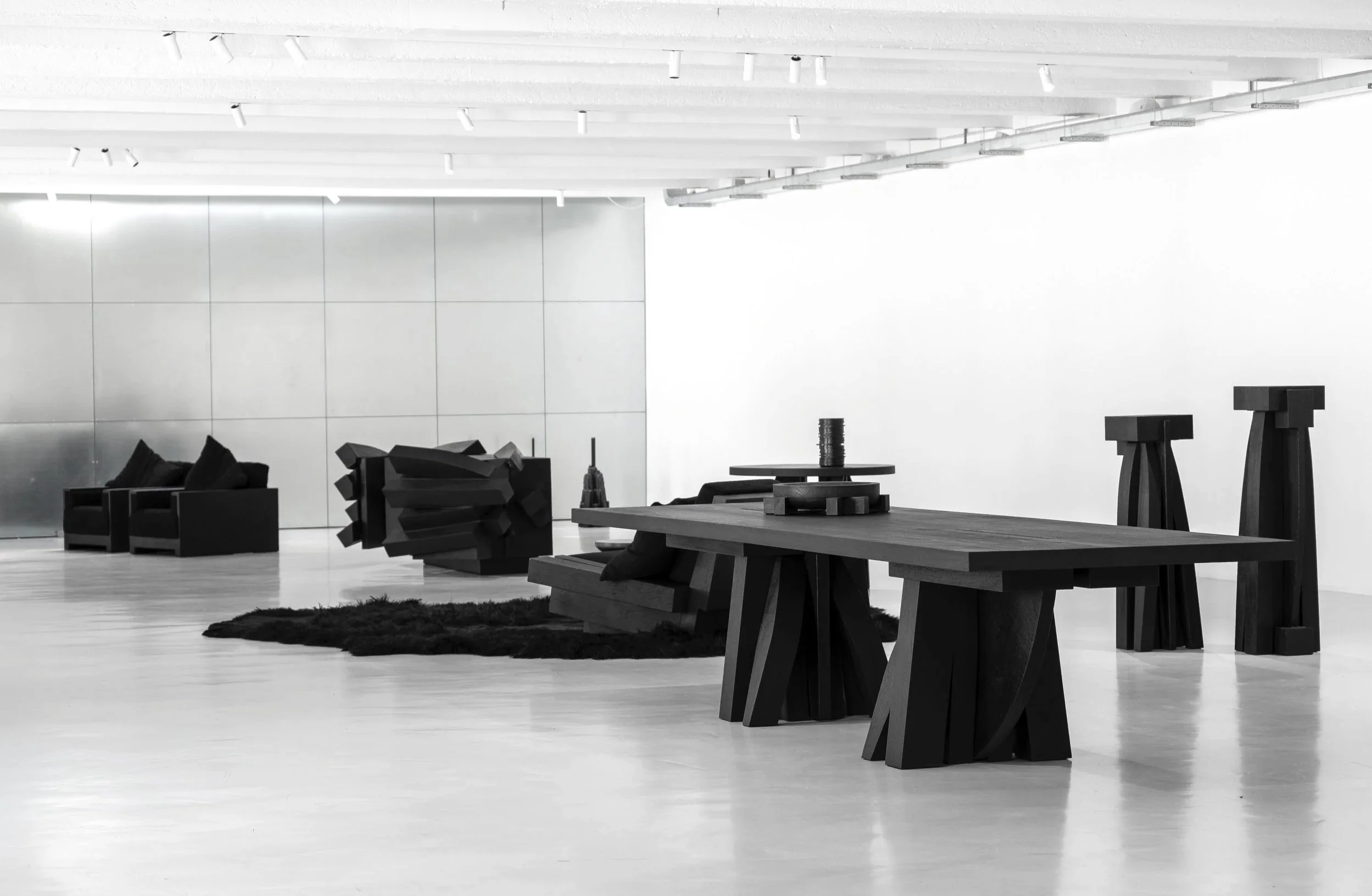 Minimalist modern interior with black geometric furniture including chairs, a large wooden table, and abstract sculptures, in a spacious white room.