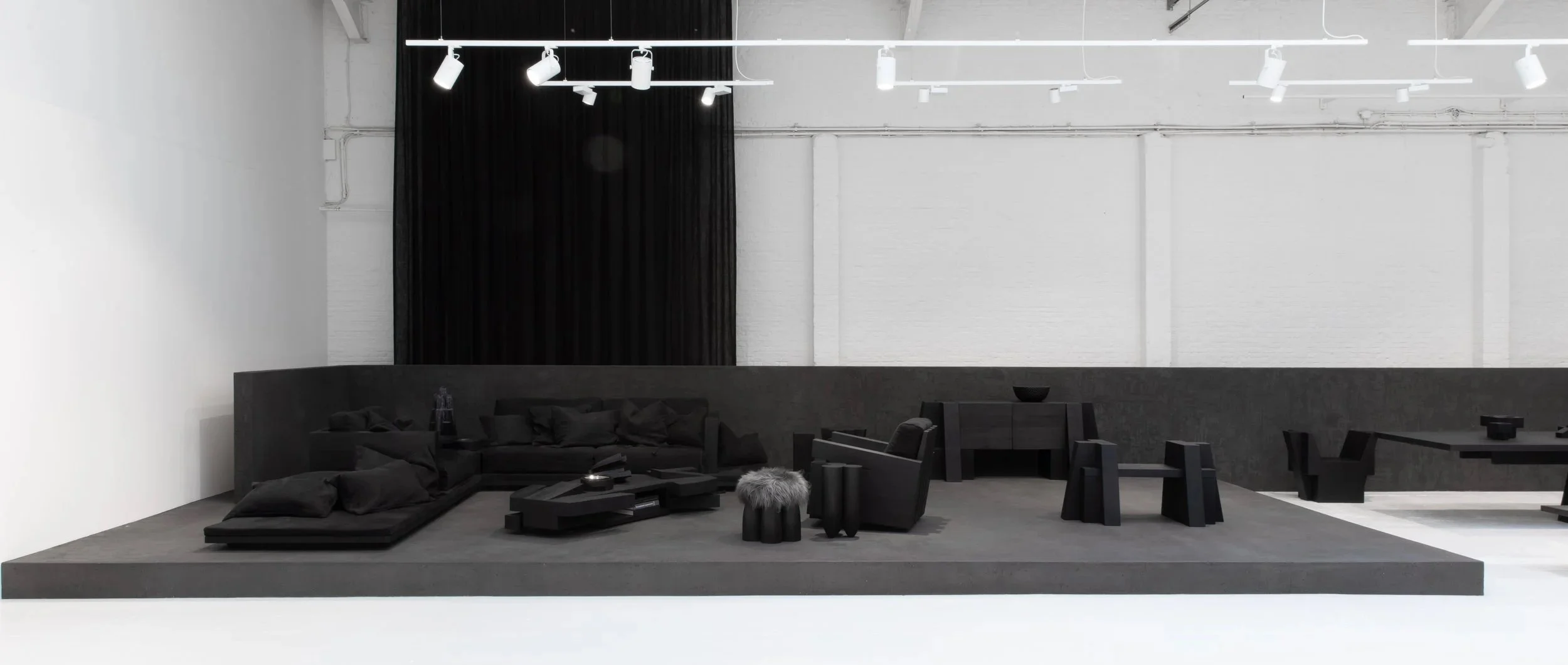 Minimalist interior with black furniture on a raised gray platform, black couches, chairs, stools, a table, and a black curtain in the background, with white walls and ceiling lights.