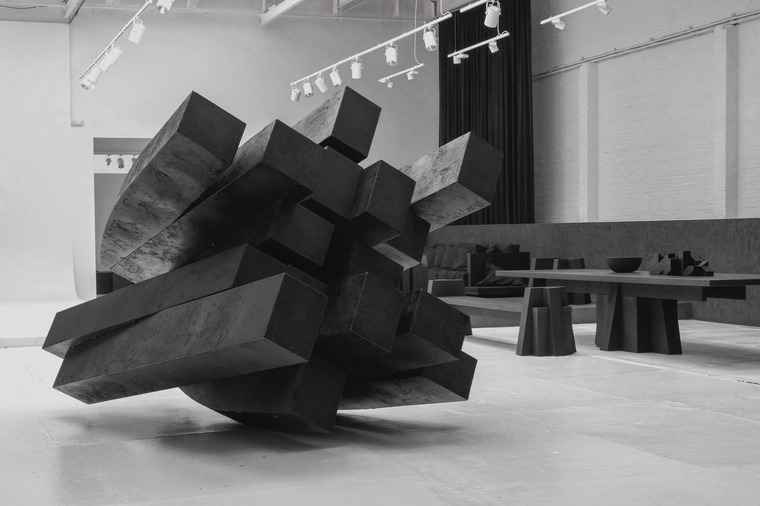 Modern, minimalist interior with black furniture including two lounge chairs with cushions, a large wooden dining table with three tall chairs, and several abstract black sculptures. The room has white walls and ceiling, with track lighting and a glass wall in the background.