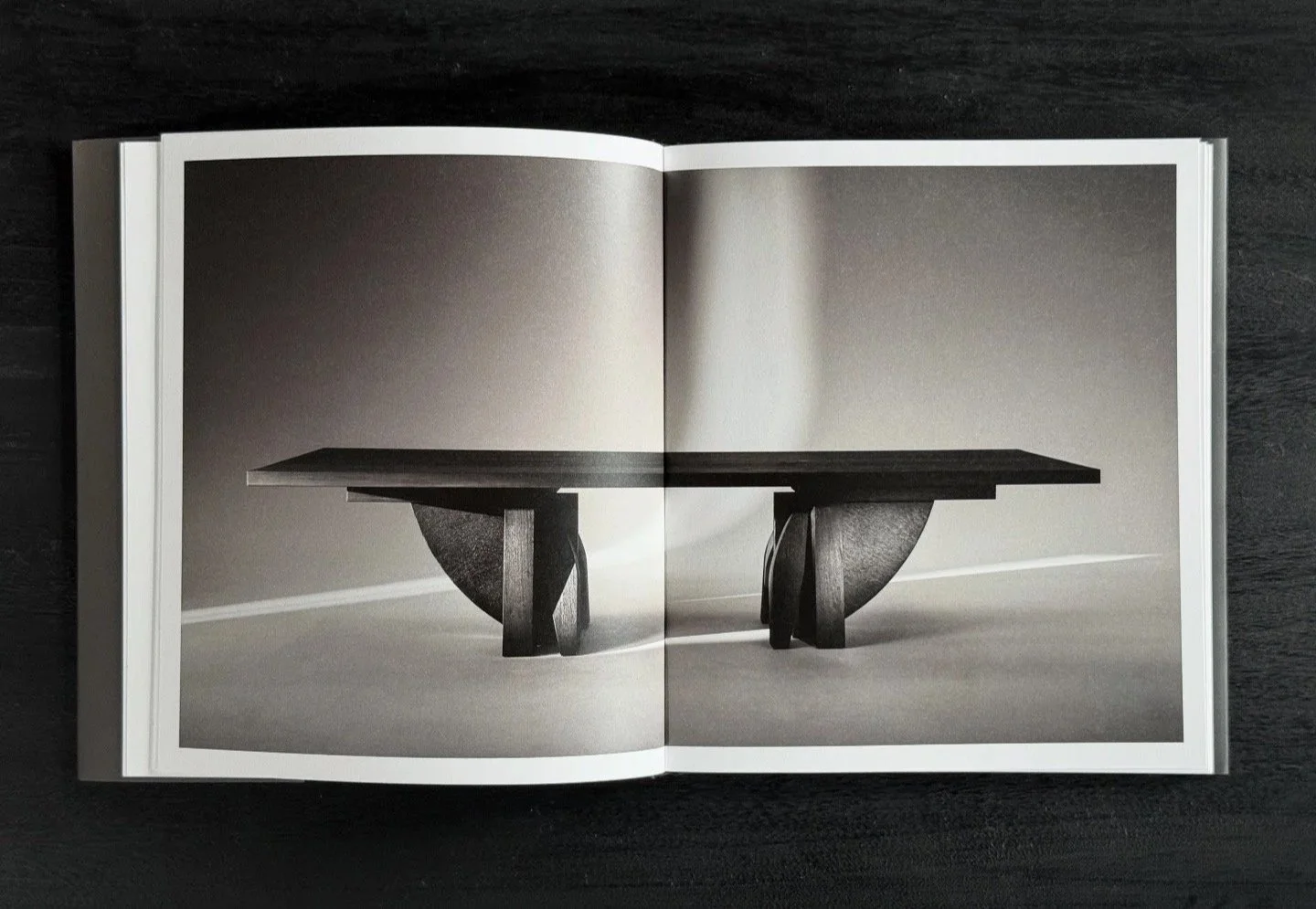Open book displaying a black-and-white photograph of a modern wooden table with a rectangular top and unique curved supports, set against a plain background.