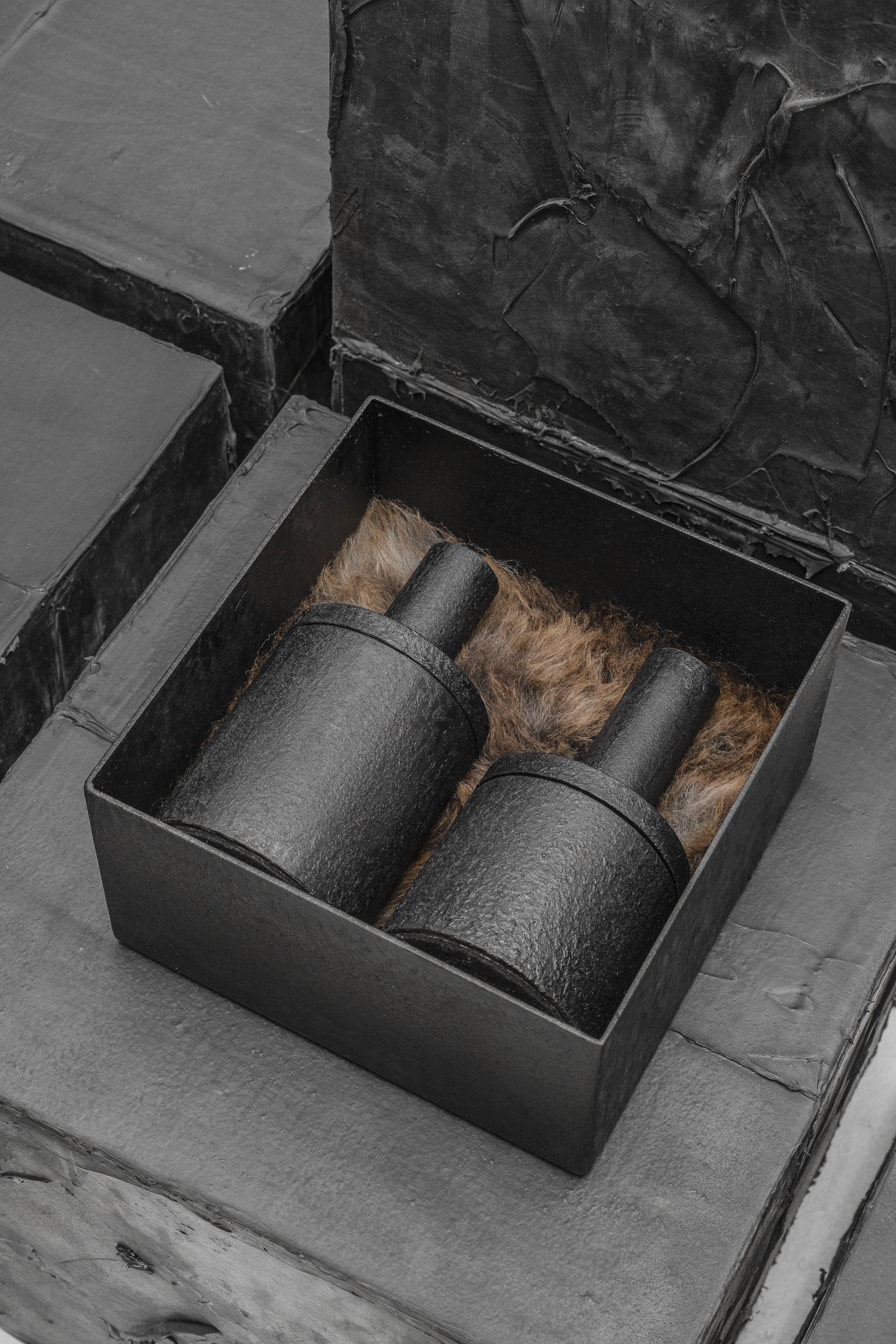 Two black cylindrical metal objects placed in a black box with a brown furry lining, on a gray surface with black textured material in the background.