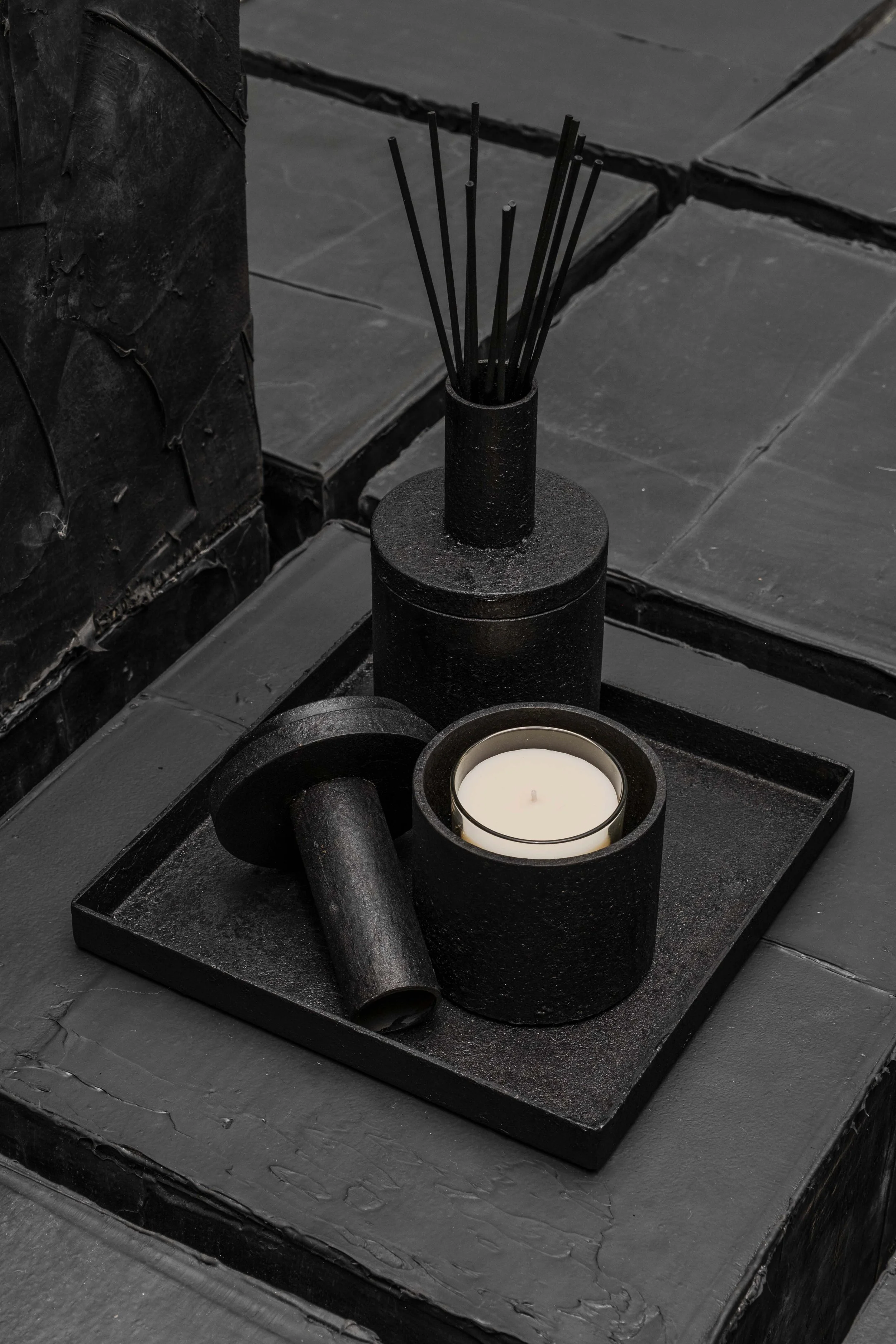 Black and white photo of a decorative tray with a candle, a cylindrical container, and a reed diffuser on a textured dark surface.