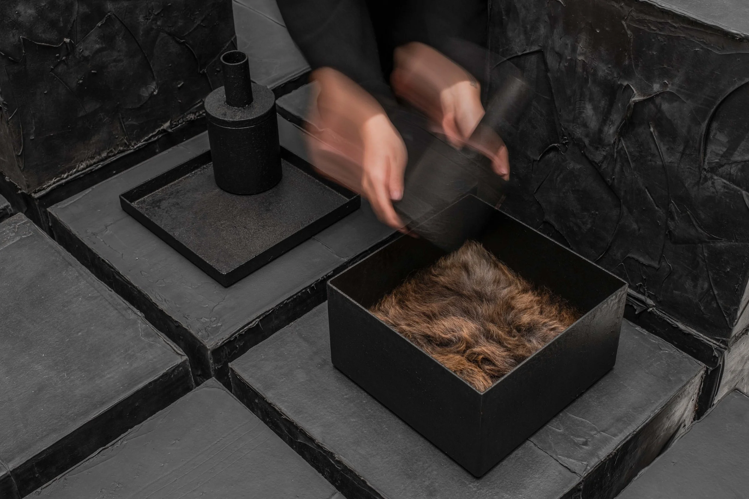 Person handling a black box of fur in a dark, industrial setting with black tiled flooring.